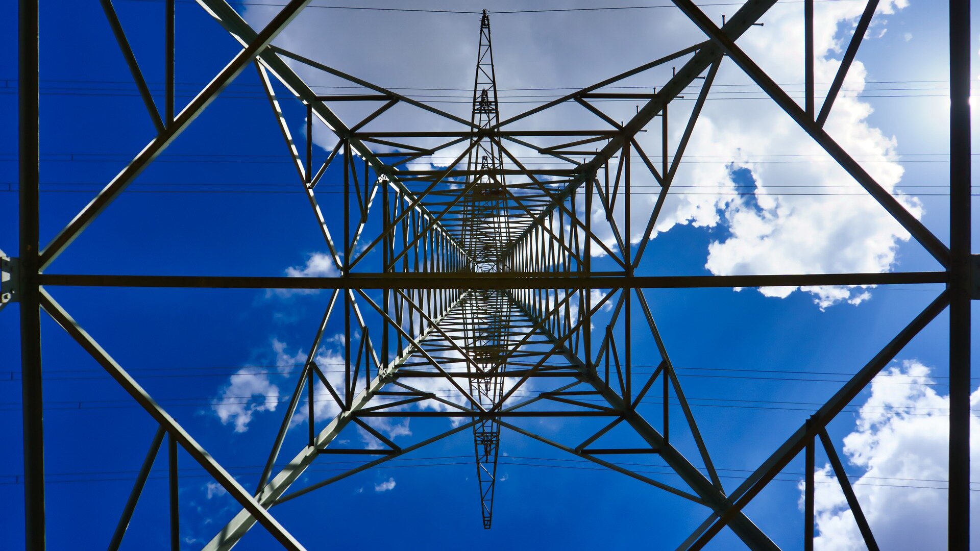Na zdjęciu są czarne pręty konstrukcji słupa energetycznego. Konstrukcja jest symetryczna. W poprzek słupa w jego górnym odcinku jest pozioma konstrukcja. W tle niebieskie niebo z białymi chmurami. 
