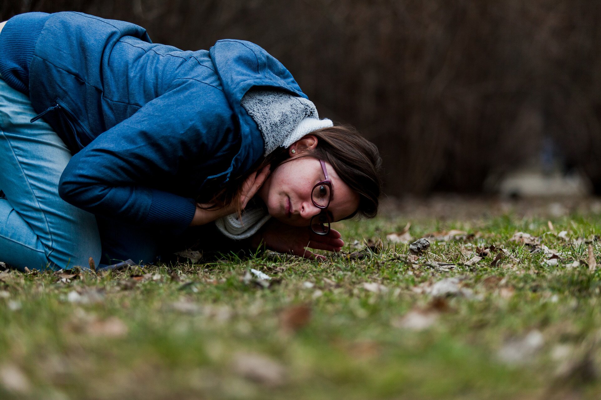 Zdjęcie kobietę w okularach, ubraną w niebieską kurtkę nasłuchującą zwierząt z uchem przy ziemi. Osoba jest pochylona nad ziemią ponieważ przy samej ziemi dźwięki te słyszalne są najlepiej.