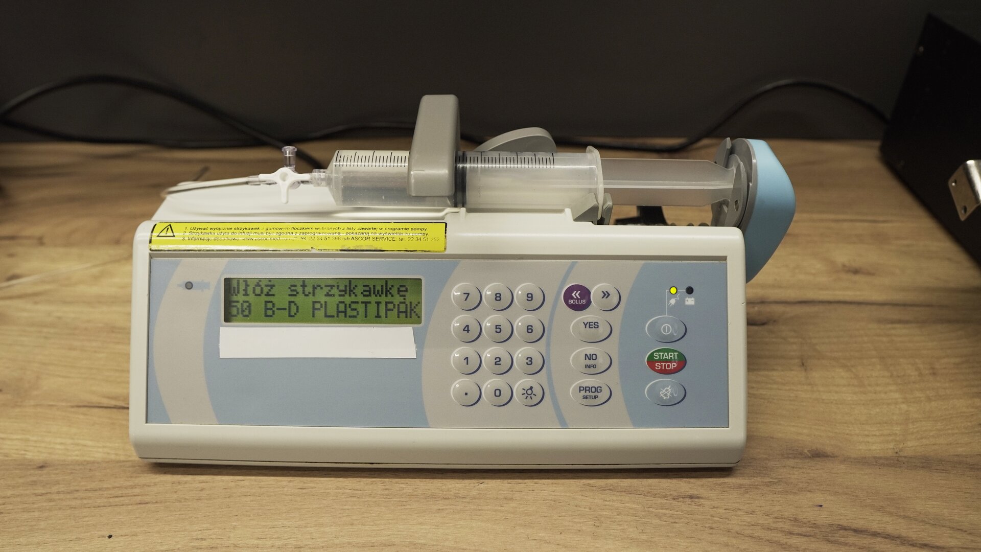 Na zdjęciu strzykawka wypełniona wodą znajduje się w ramieniu pompy infuzyjnej. Na ekranie aparatu wyświetlana jest informacja: "Włóż strzykawką 50 B‑D PLASTIPAK".