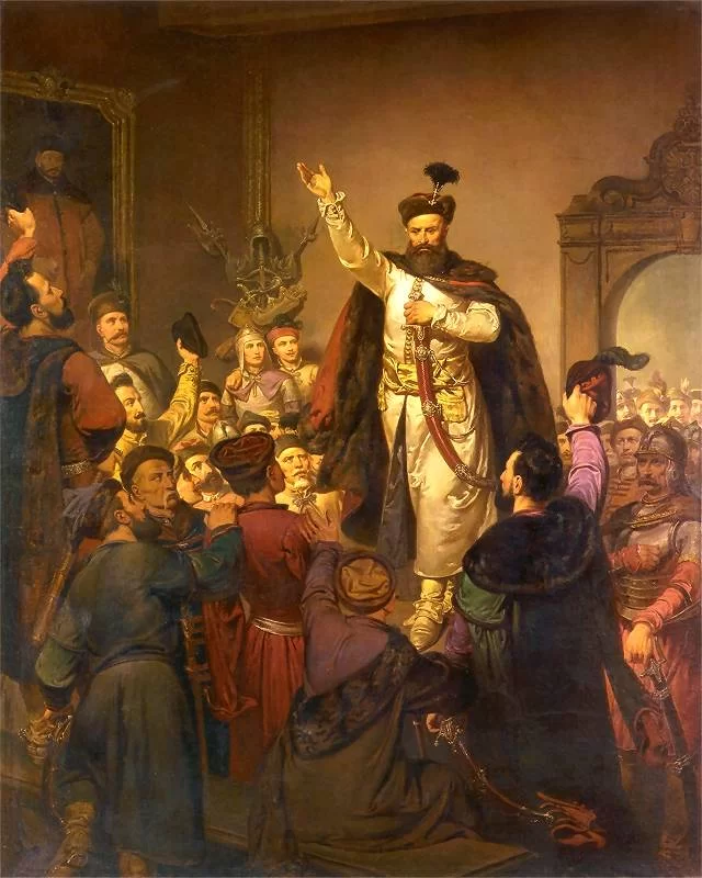 Kliknij, aby powiększyć. Na obrazie ludzie we wnętrzu pomieszczenia. w centralnej części na podwyższeniu mężczyzna w średnim wieku. Ma on gęstą ciemną brodę i wąsy. Na głowie ma czapkę z ozdobnym pomponem. Jego prawa ręka jest uniesiona w górę. W lewej dłoni trzyma szablę skierowaną w dół. Biodra obwiązane są szerokim pasem, a na ramionach jest płaszcz. Wokół mężczyzny stoi tłum mężczyzn. Niektórzy podnieśli czapki do góry. Po prawej stronie stoi żołnierz w pancerzu. W tle widać tłum ludzi w wejściu. Po lewej stronie na ścianie wisi obraz.