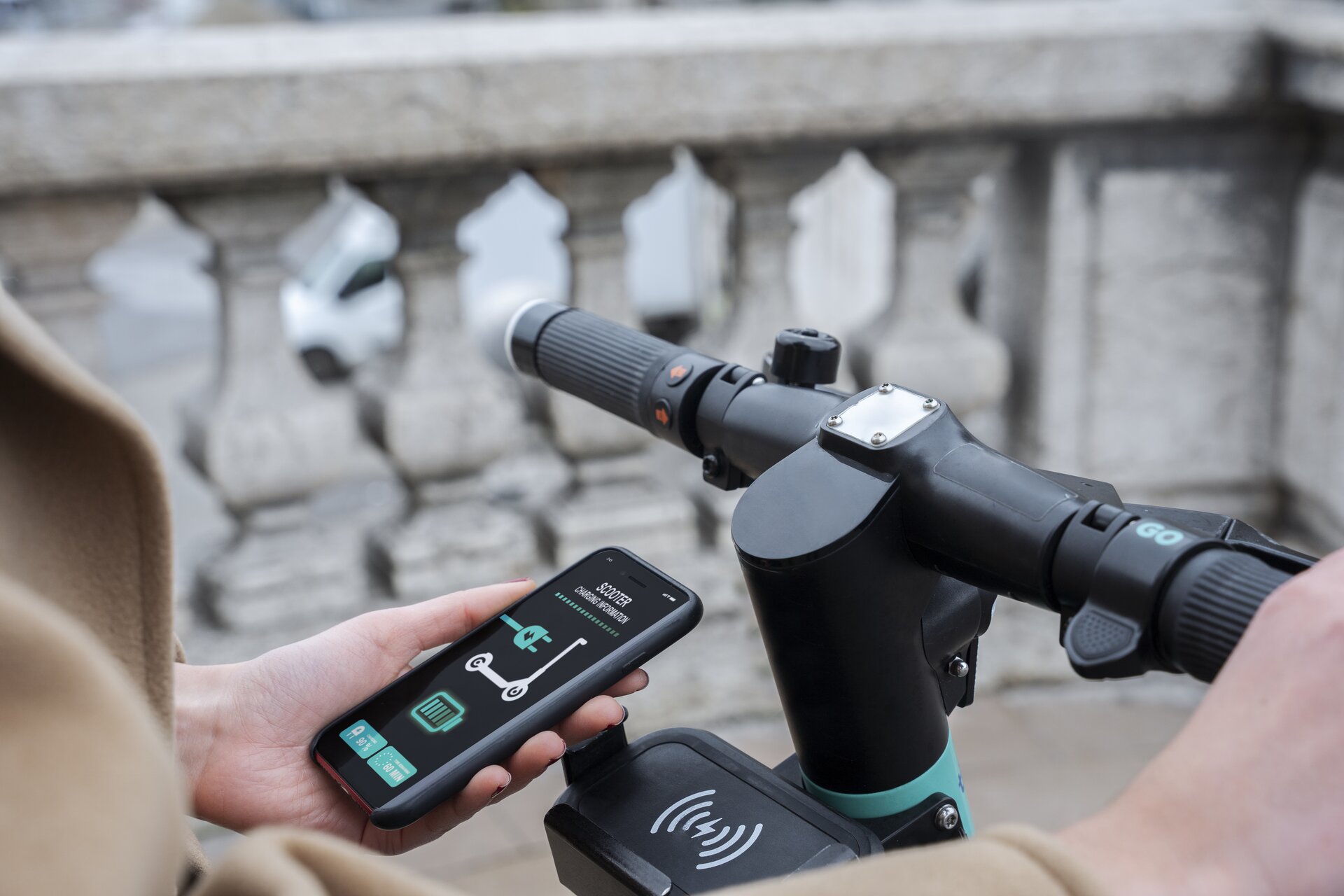 Zdjęcie przedstawia kierownicę hulajnogi elektrycznej. Widoczna również dłoń, która trzyma baterię ładującą hulajnogę. Na ekranie wyświetlacza widoczna ikona hulajnogi i wtyczki do gniazda elektrycznego oraz poziom naładowania. 