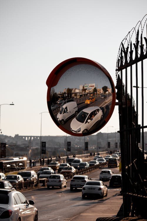 Zdjęcie obrazu powstającego w zwierciadle kulistym. Lustro zawieszone jest na ogrodzeniu. W odbiciu autostradę oraz budynki. Za lustrem widoczne są samochody poruszające się po autostradzie.