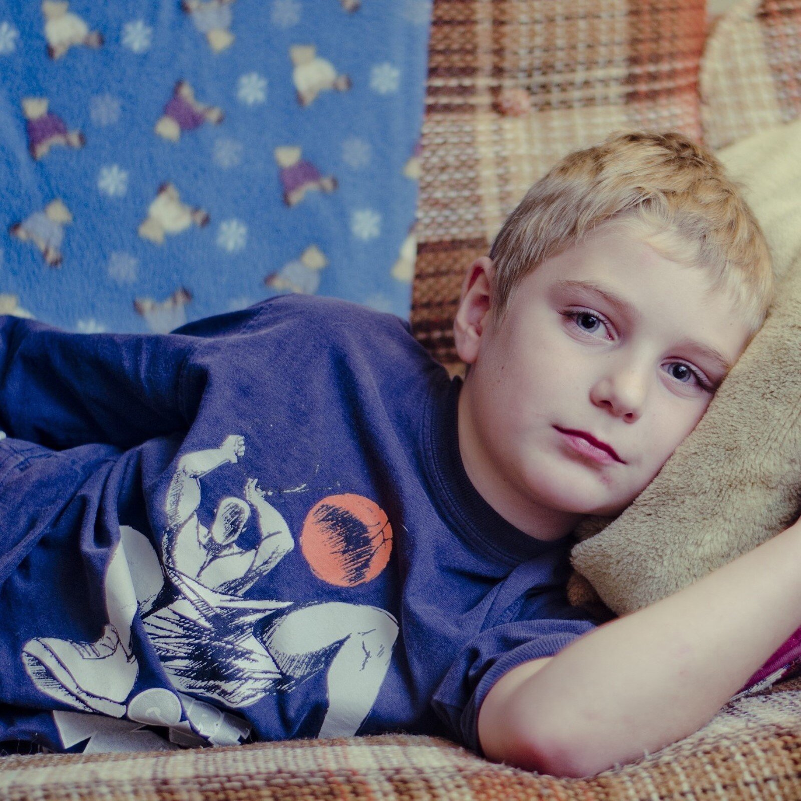 Zdjęcie przedstawia leżącego na kanapie chłopca, w ujęciu od pasa w górę. Chłopiec ma krótkie, jasne włosy, jest ubrany w bluzkę z superbohaterem, leży na lewym boku i opiera głowę na poduszce.