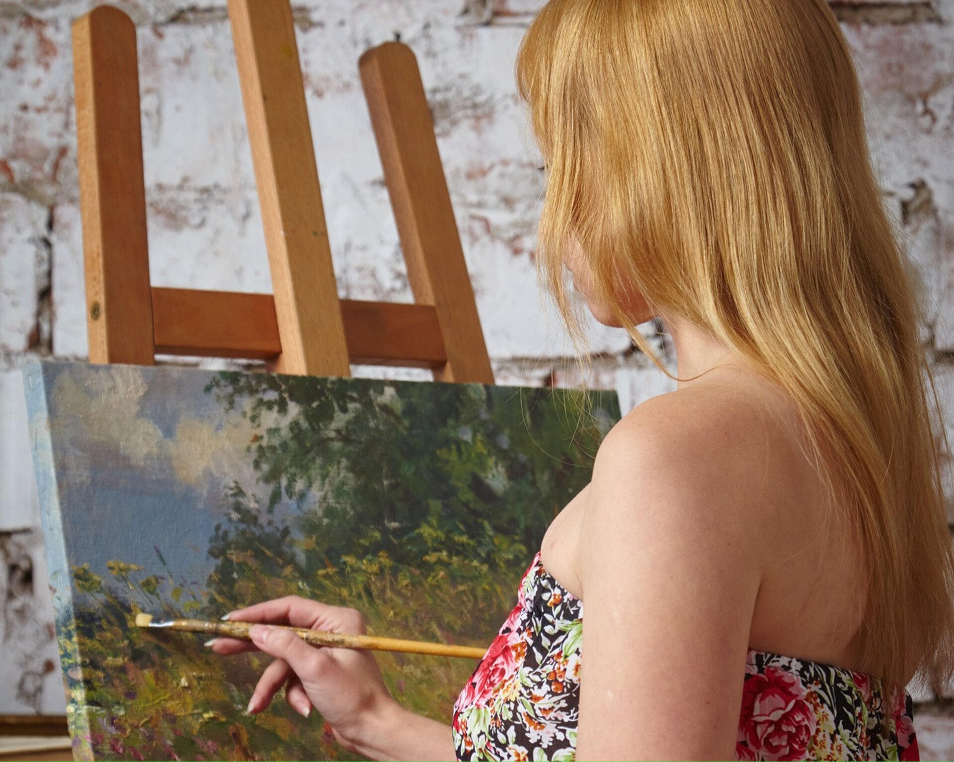 Zdjęcie przedstawia długowłosą kobietę, która stoi przy sztaludze i maluje pejzaż.