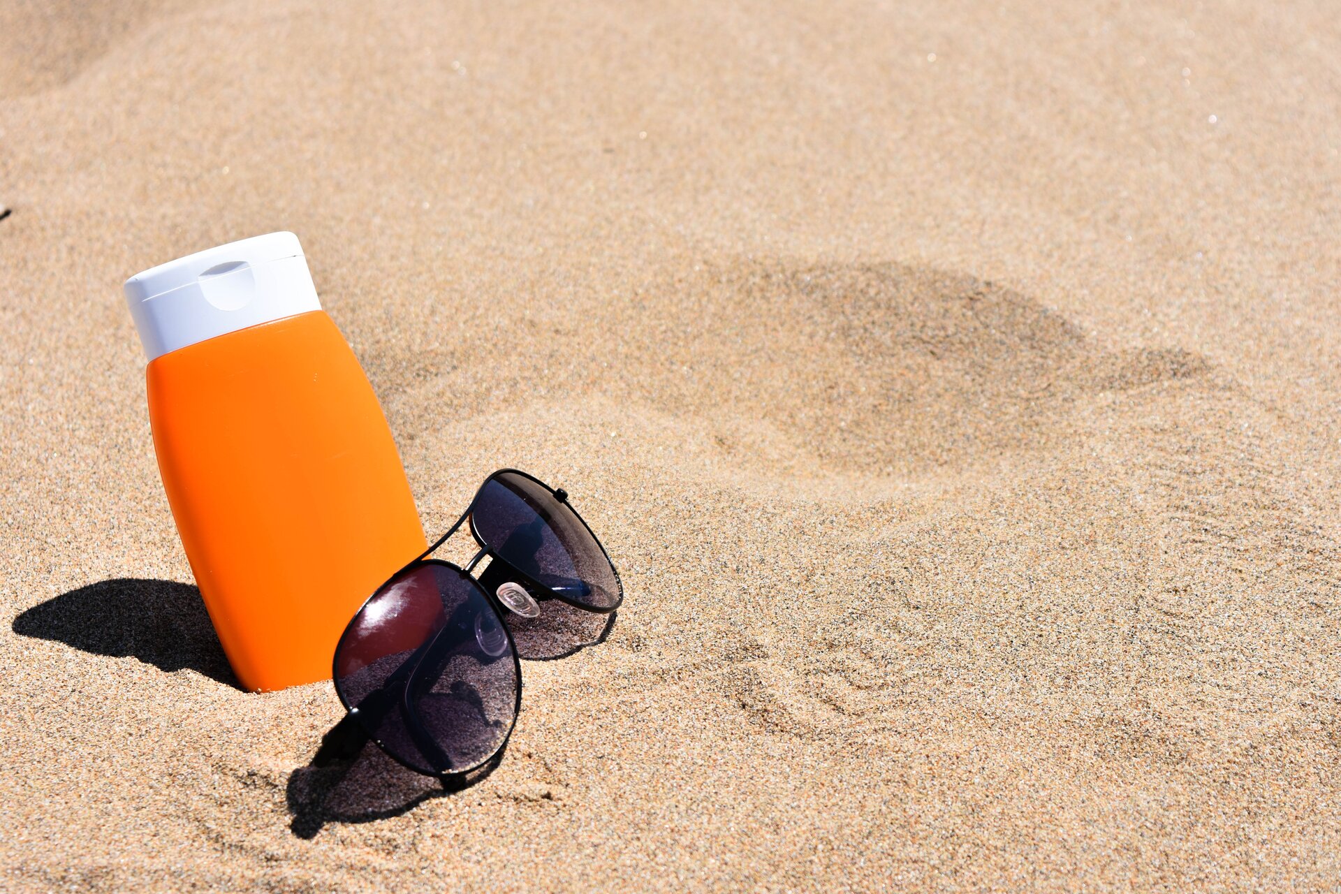 Na zdjęciu fragment plaży z drobnym złocistym piaskiem. Po lewej stronie leżą okulary słoneczne, za nimi stoi plastikowa butelka preparatu do opalania w kolorze pomarańczowym z białą nakrętką.