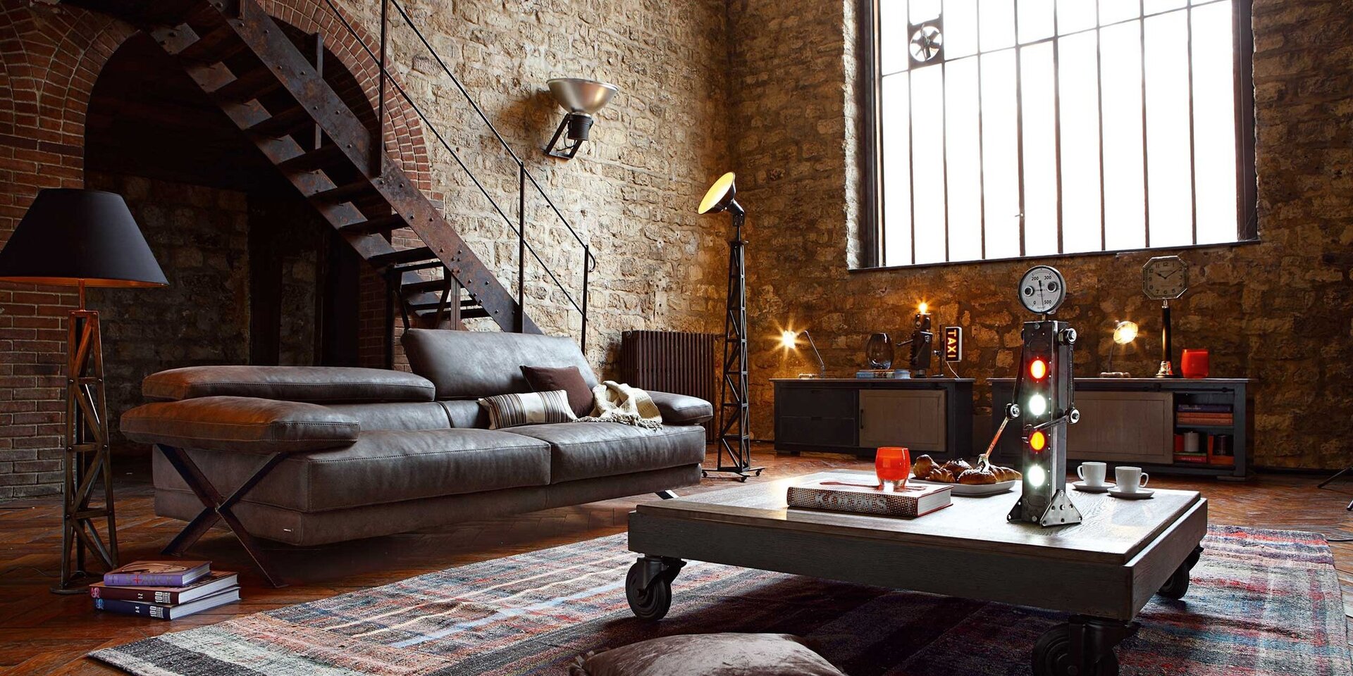 Ilustracja przedstawia pomieszczenie pokryte cegłą. Na prawej ścianie widnieje duże fabryczne okno z wbudowanym wiatrakiem w lewej jego części. Pod oknem stoją dwie komody. Obie komody są ciemne, ale posiadają one kontrastowe do ich koloru jasne drzwiczki. Na lewej komodzie jest lampka, szklany wazon na książkach, robot z żarówką na głowie oraz podświetlany znak taxi. Na prawej komodzie zaś stoi lampka, industrialny zegar oraz czerwony wazon. Drzwi prawej komody są otwarte. W środku tej komody są książki, puszki i kartony. Na lewej ścianie widać duży metalowy, rdzawy grzejnik. Zaraz przy nim znajdują się  mosiężne metalowe, rdzawe schody. Na ścianie za schodami widnieje ceglany łuk, a w nim dalszą, pustą część pomieszczenia. Przed schodami stoi duża skórzana kanapa na metalowych wspornikach. Po lewej stronie kanapy dostrzegamy lampę na metalowym stojaku. Lampa ma czarny klosz. Pod lampą leżą 3 książki. Przed sofą na dywanie stoi stolik. Stolik wspiera się o ruchome kółka. Blat stołu wykonany jest z drewna. Na stole leży książka, na której postawiona jest świeczka. Widać tam również talerz z wypiekami, dwie filiżanki z talerzykami oraz robota, z licznikiem z ruchomymi oczkami zamiast głowy. Tors robota posiada cztery lampki: czerwoną, zieloną, czerwoną, zieloną. Po prawej stronie sofy stoi wysoka czarna lampa na metalowym stelażu.