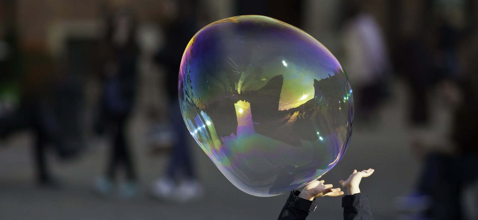 Zdjęcie przedstawia unoszącą się w powietrzu ogromną bańkę mydlaną, która mieni się kolorami tęczy. Próbują jej dosięgnąć dziecięce rączki. W tle, które jest niewyraźne, widać przechodzące osoby.