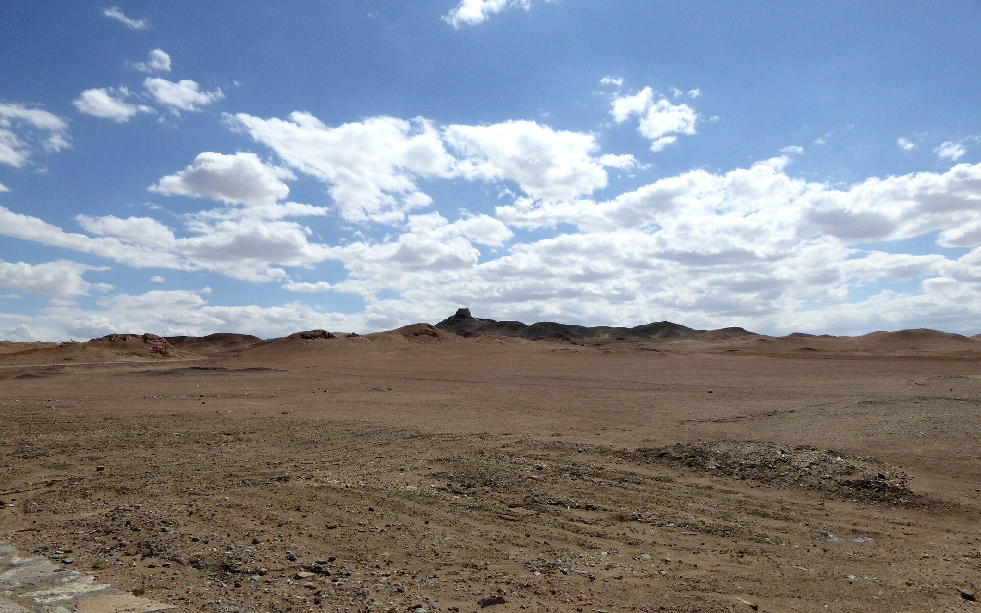 Zdjęcie przedstawia Pustynię Gobi w Chinach. Pustynny krajobraz utworzony jest z rozległej płaszczyzny zbudowanej z brązowej ziemi. Na horyzoncie łączy się z błękitnym niebem z białymi obłokami.