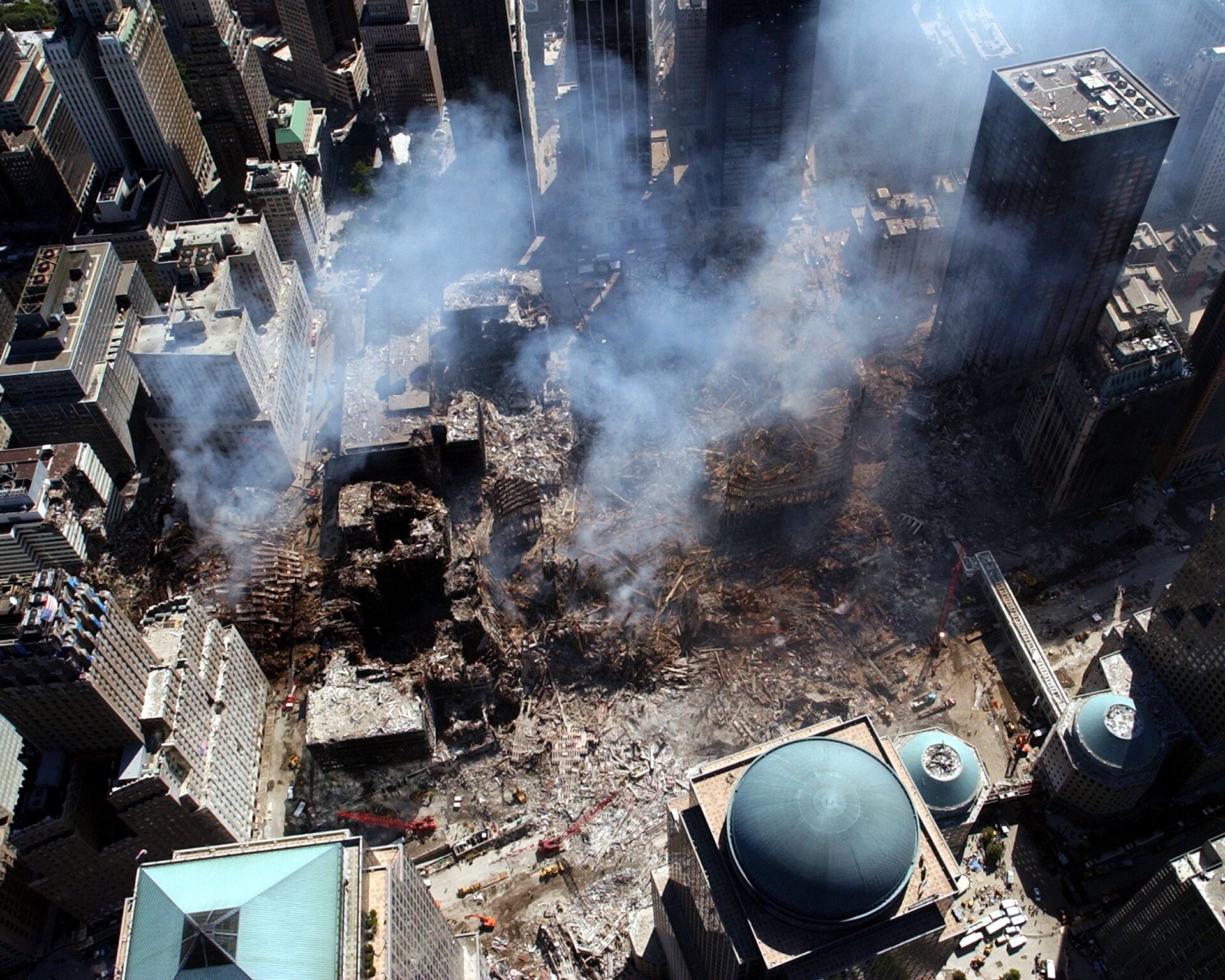 Zdjęcie przedstawia widok z góry na zgliszcza dwóch wież World Trade Center.  Widać ogrom zniszczeń. Dookoła stoją wieżowce. 