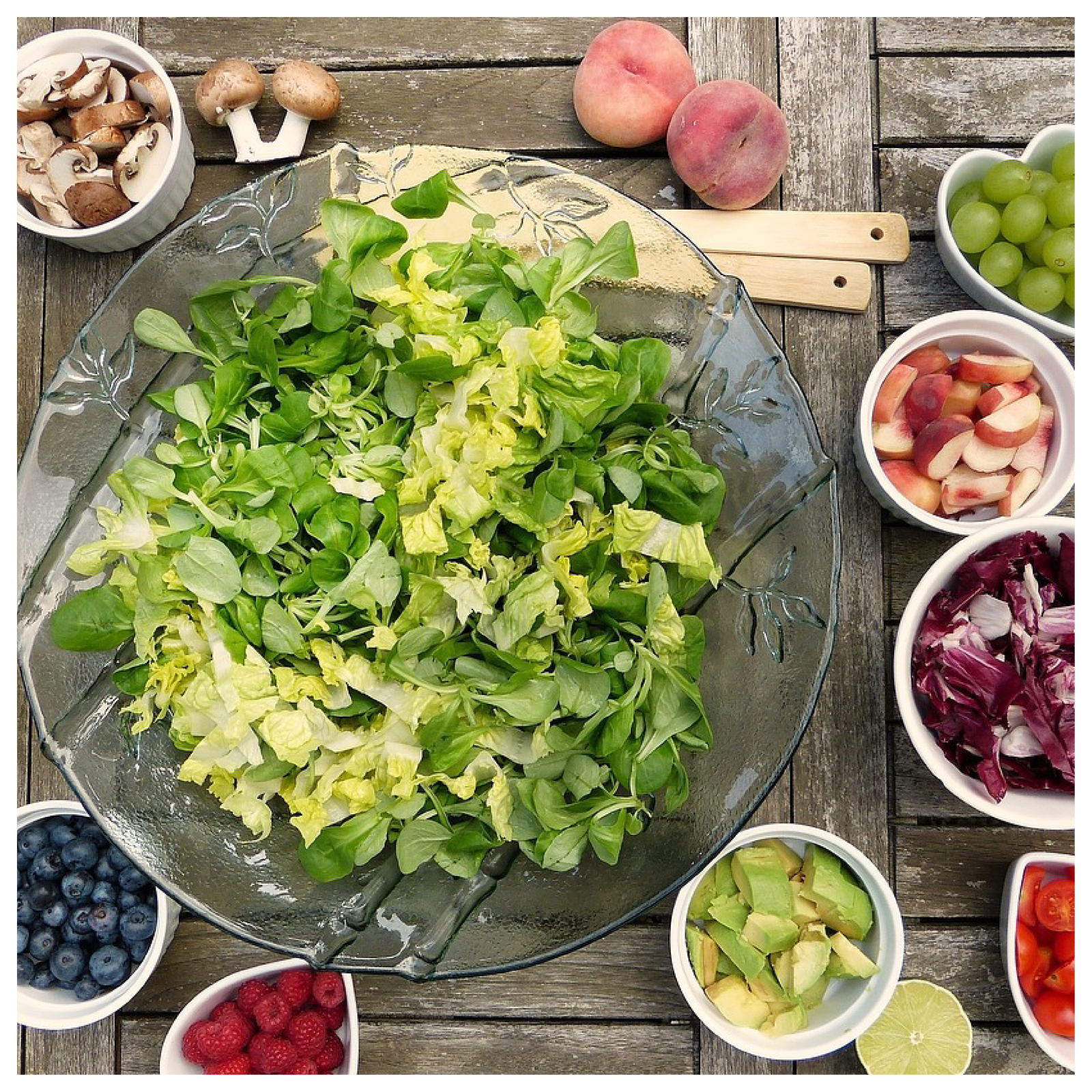 Na zdjęciu widoczny jest stół, na którym leżą miski z różnymi produktami spożywczymi. Po środku leży największa miska z sałatą. Obok stoją mniejsze miseczki, w których znajdują się pokrojone: pieczarki, winogrona, orzechy, awokado, truskawki, pomidorki koktajlowe, borówki i czerwona sałata.