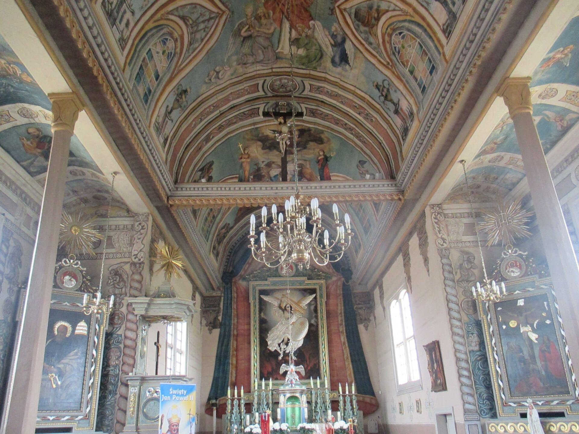 Zdjęcie przedstawia wnętrze kościoła. Zarówno ściany, jak i sufit są ozdobione malowidłami przedstawiającymi sceny biblijne. Widoczny w centralnej części zdjęcia ołtarz jest ozdobiony dużym obrazem, przedstawiającym anioła. Przed obrazem znajduje się zielone tabernakulum, a także dużo świec.