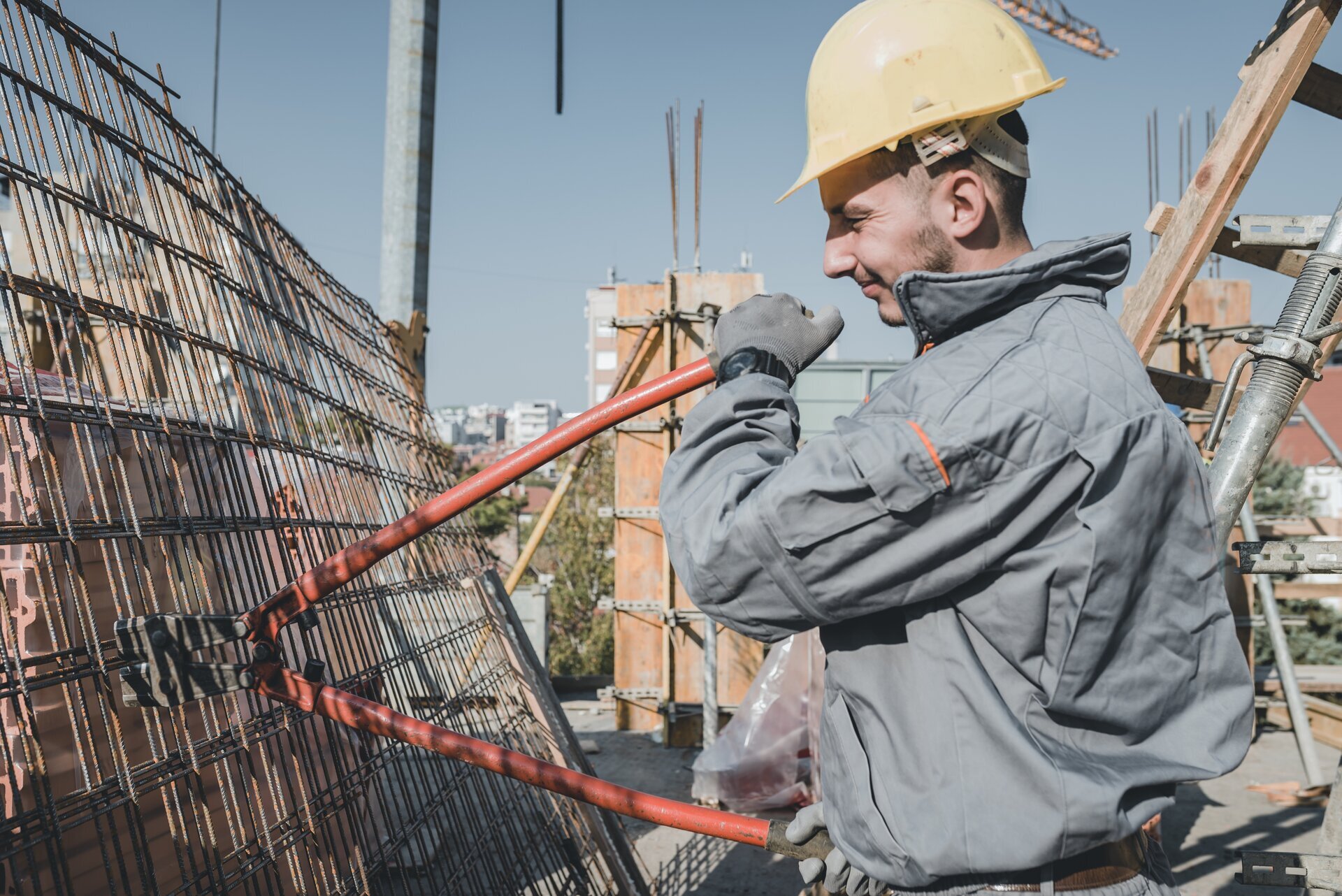 Zdjęcie przedstawia nożyce zbrojarskie ręczne w kolorze czerwonym, którymi robotnik w żółtym kasku przecina stalową siatkę.
