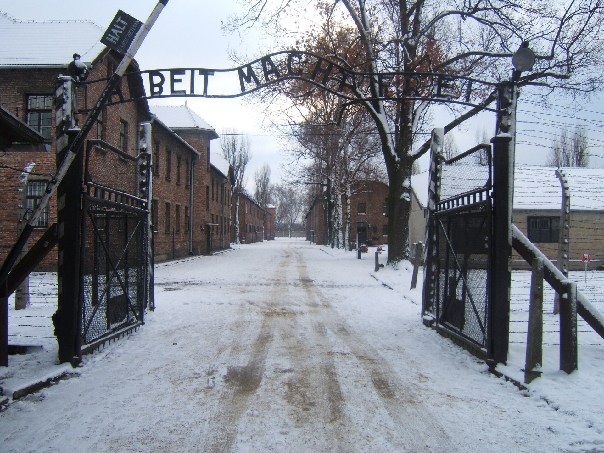 Zdjęcie przedstawia bramę wjazdową do Auschwitz. Widnieje na niej niemiecki napis: Arbeit macht frei, który oznacza "Praca czyni wolnym". Za bramą widać ceglane budynki ustawione po obu stronach drogi. Płot jest zrobiony z drutu kolczastego.