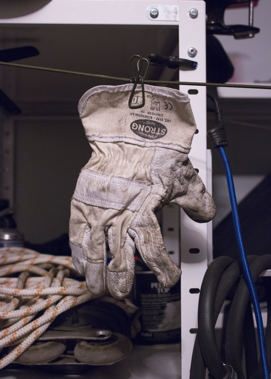 Ilustracja przedstawia fotografię „Rękawiczka”. Na zdjęciu ukazana jest podniszczona rękawica robocza zawieszona na zielonej linie. W tle znajduje się fragment regału z półkami, na których leżą liny, puszki, wąż gumowy, niebieski kabel oraz metalowe narzędzia.