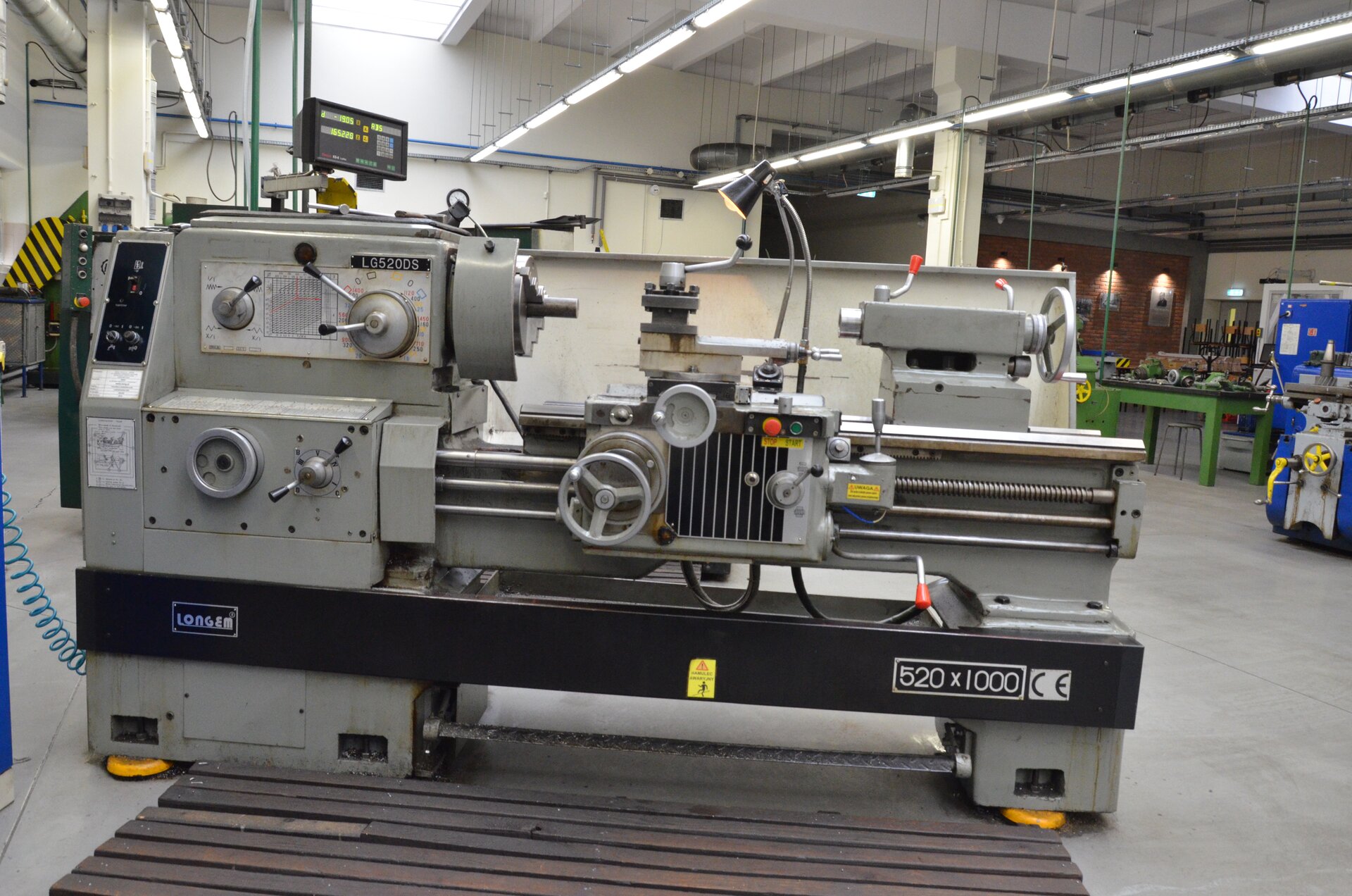 Ilustracja przedstawia tokarkę w warsztacie. Tokarka zbudowana jest z łoża, wrzeciona, konika, skrzynki prędkości obrotowych, skrzynki posuwów, skrzynki suportowej, imaka nożowego, śruby pociągowej, wałka pociągowego, wałka sterującego, gitary oraz wyświetlacza z odczytem cyfrowym położenia. 