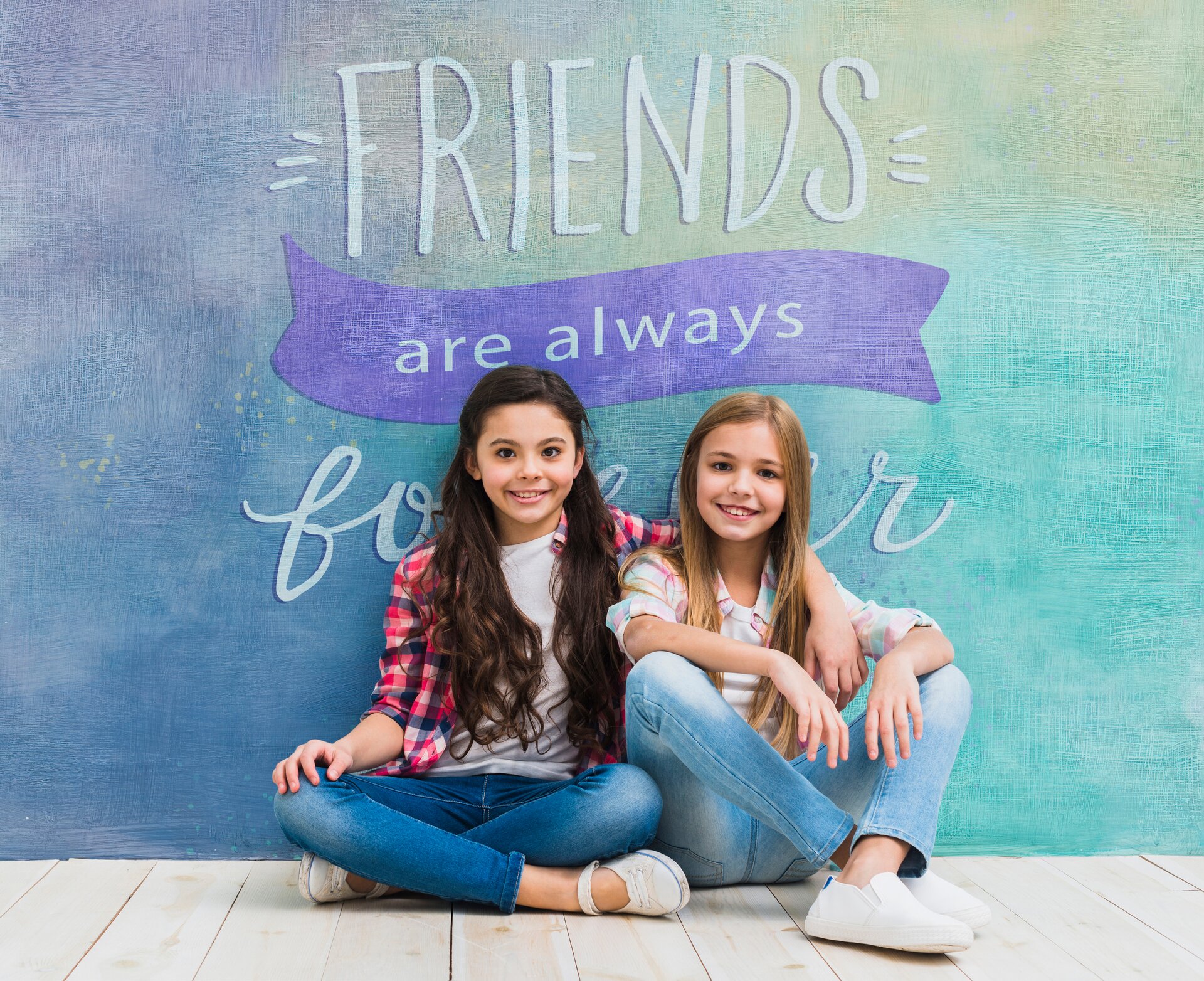 Zdjęcie przedstawia dwie dziewczynki w wieku szkolnym siedzące po turecku pod kolorową ścianą, na której widoczny jest napis w języku angielskim: FRIENDS ARE ALWAYS . Dziewczynki opierając się o ścianę zasłaniają  słowo:   FOREVER. 