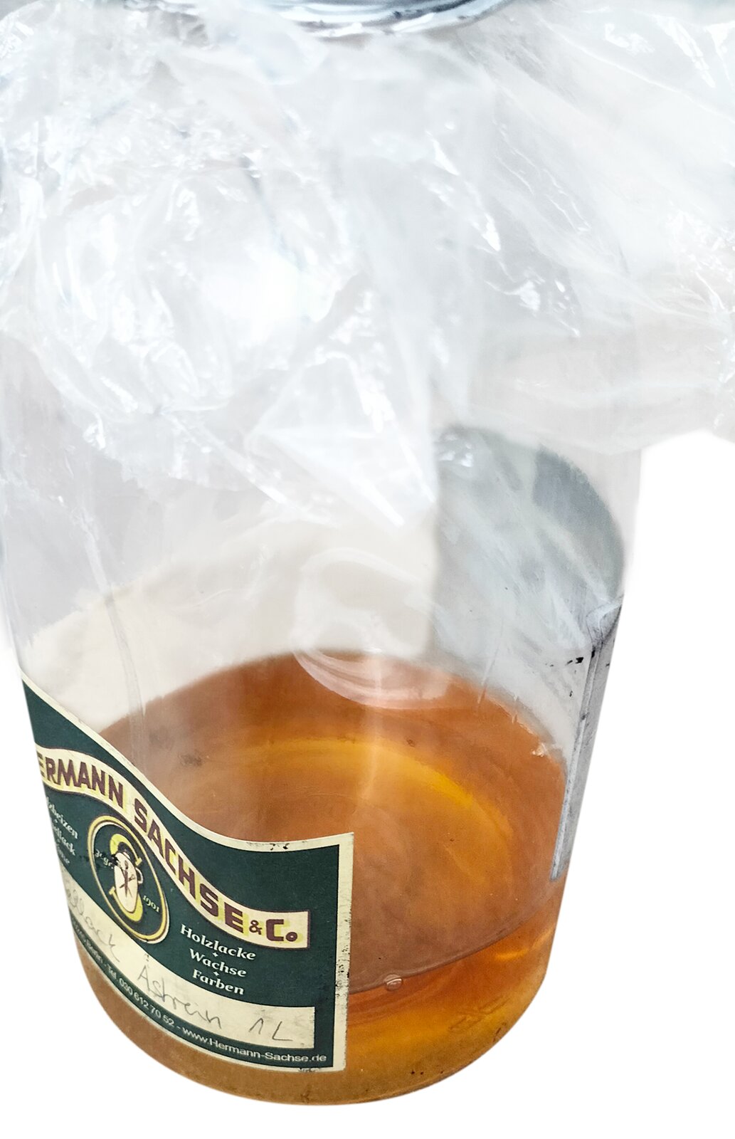 Zdjęcie przedstawia politurę. Widoczny jest przeźroczysty pojemnik, z nałożoną przeźroczystą folią w górnej części, w środku którego znajduje się bursztynowy płyn.