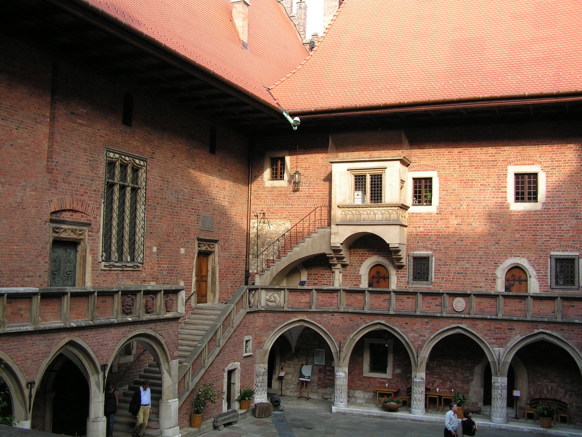 Zdjęcie przedstawia najstarszy budynek Uniwersytetu Jagiellońskiego tak zwane Collegium Maius. Fotografia została wykonana wewnątrz zabudowań, z perspektywy balkonu, z którego widok rozciąga się na dziedziniec z krużgankami. Podcienia o strzelistych łukach, wsparte są na zdobionej kolumnadzie.
