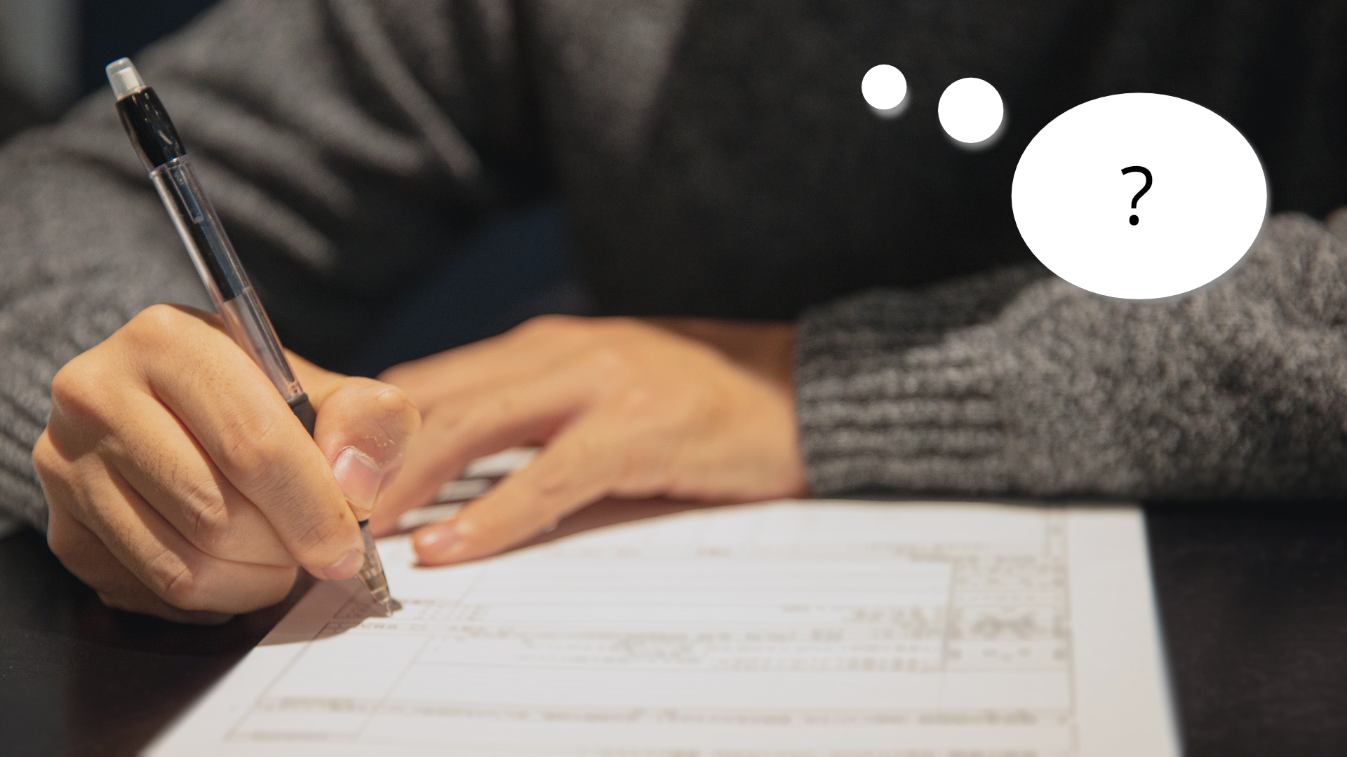 Zdjęcie przedstawia ręce osoby trzymającej w ręku długopis i wypełniającej formularz. Obok w dymku znajduje się znak zapytania.