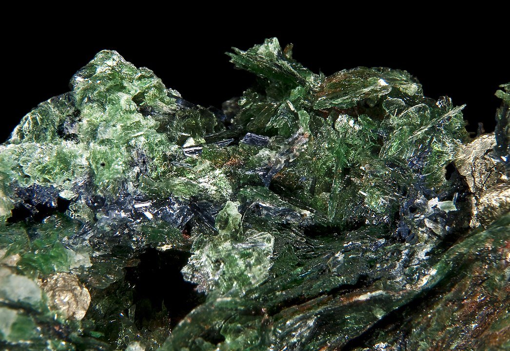 Zdjęcie przedstawia muskowit. Jest to ciemnozielona skała, która charakteryzuje się połyskiem i krystaliczną budową. Ma nieregularny kształt, miejscami posiada jasnoszare plamy.