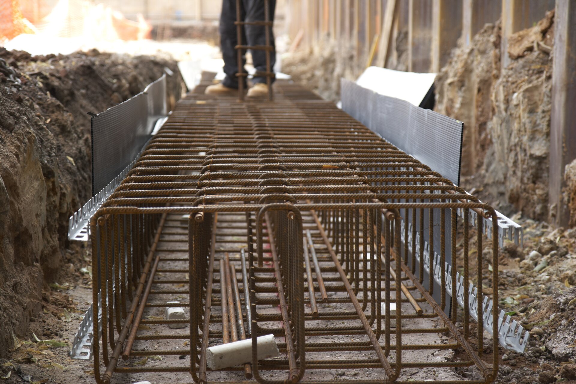 Zdjęcie przedstawia zbrojenie fundamentu. Na drugim planie widać nogi robotnika trzymającego stalowy pręt.