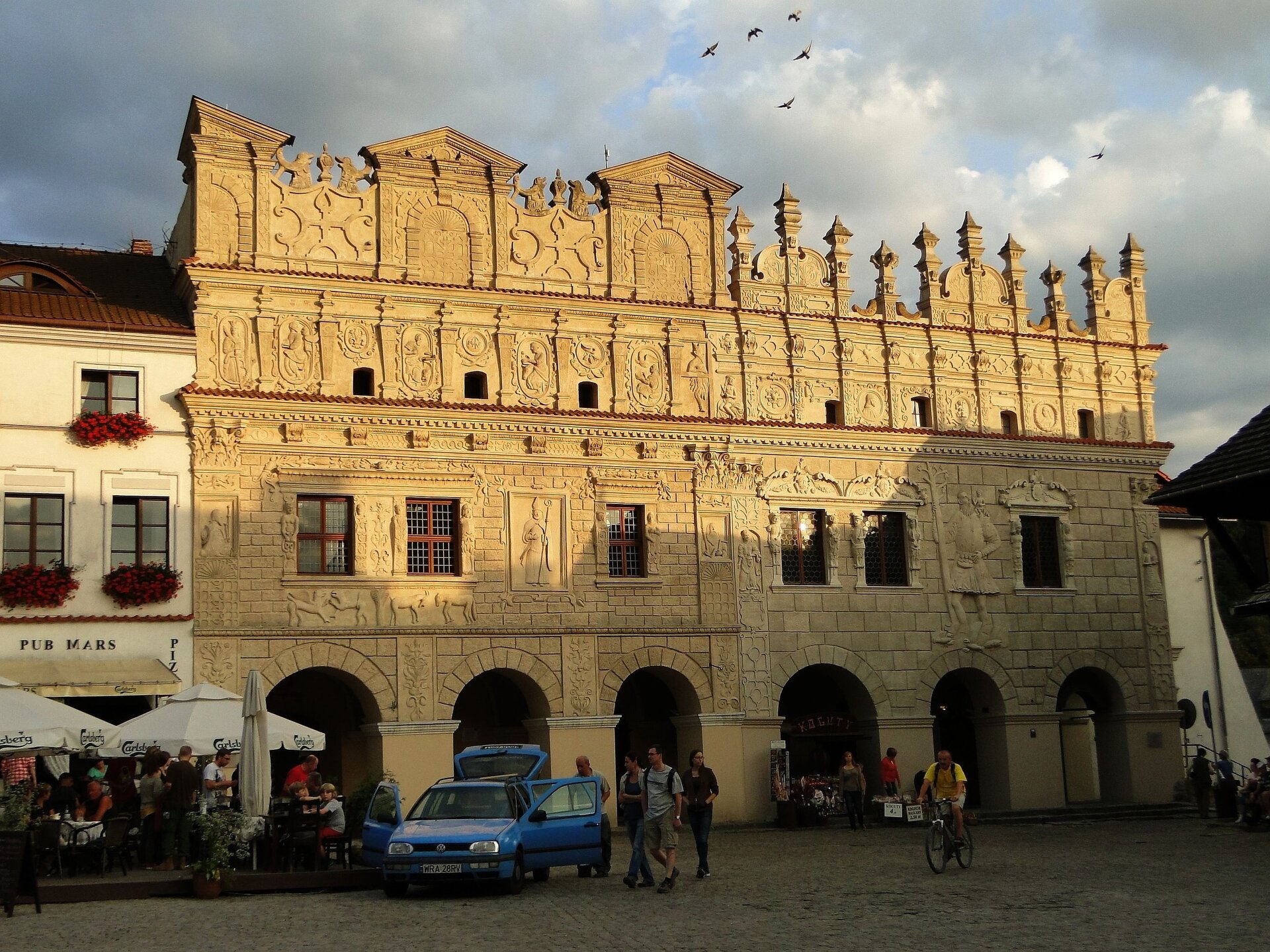 Ilustracja przedstawia Kamienice Przybyłów w Kazimierzu Dolnym. Święty Mikołaj w lewej ręce trzyma pastorał. Święty Krzysztof trzyma w ręku drzewo. Na ramieniu niesie błogosławiącego Chrystusa, przeprawiając Go przez rzekę. Parter zajmują podcienia, do których prowadzą trzy półkoliście zamknięte arkady. Scena walki gryfa z lwem. Scena walki kozła z lisem. Judyta z głową Holofernesa. Salome z głową Jana Chrzciciela. W dolnym pasie attyki sceny męczeństwa: Chrystus w cierniowej koronie, przedstawienie profilowe Chrystusa, Chrystus z krzyżem. Dolny pas attyki z przedstawieniami (od lewej): św. Katarzyny, Ewangelistów: Św. Mateusza, Św. Marka, Św. Łukasza, Św. Jana oraz wizerunkiem satyra. Attyki zajmują dwie kondygnacje i zakończone są ażurowym grzebieniem ze szczytami i sterczynami.