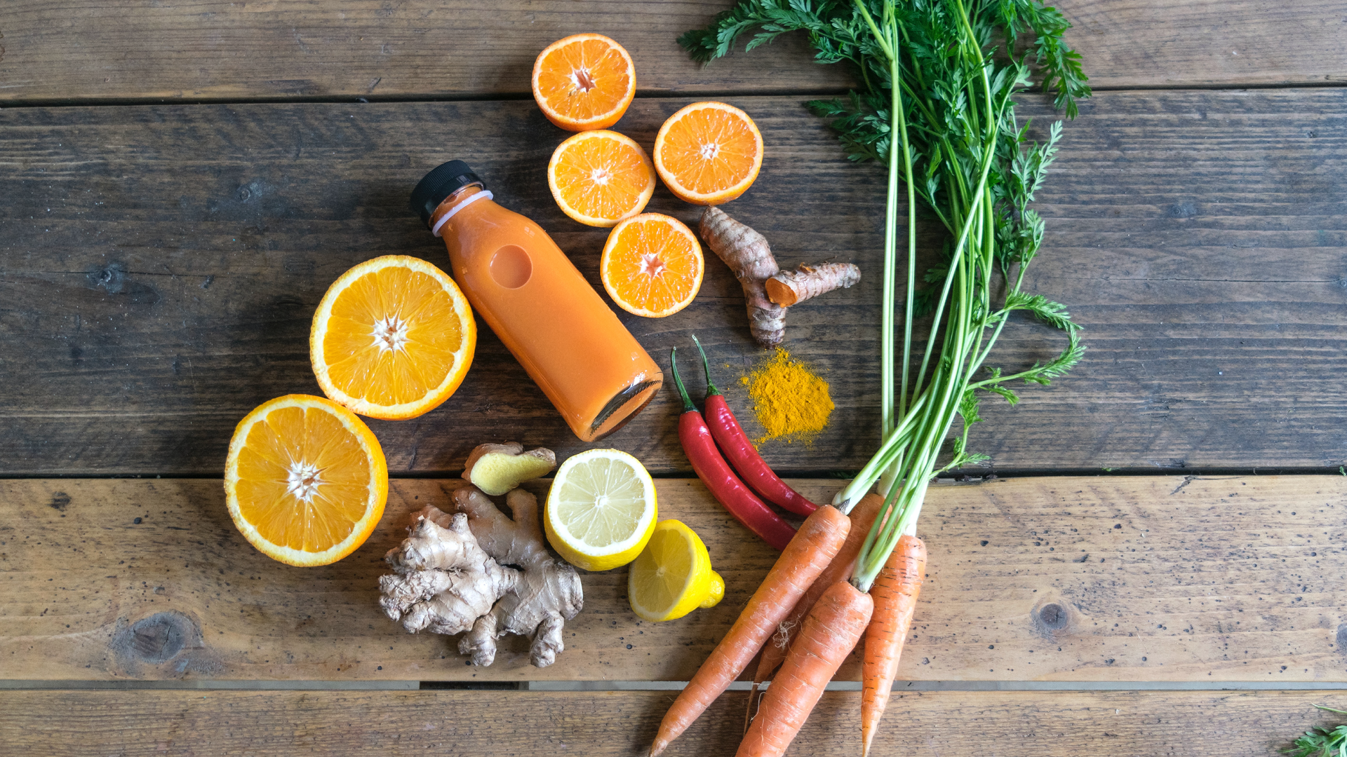 Zdjęcie przedstawia owoce, warzywa i przyprawy leżące na tle drewnianych desek. Na ilustracji znajdują się przekrojone na pół pomarańcze i cytryny, obok leży korzeń imbiru. Widoczne są również: dwie papryczki chilli, korzeń i sproszkowana kurkuma oraz pęczek marchewek z nacią. Pośrodku leży przeźroczysta butelka z sokiem.