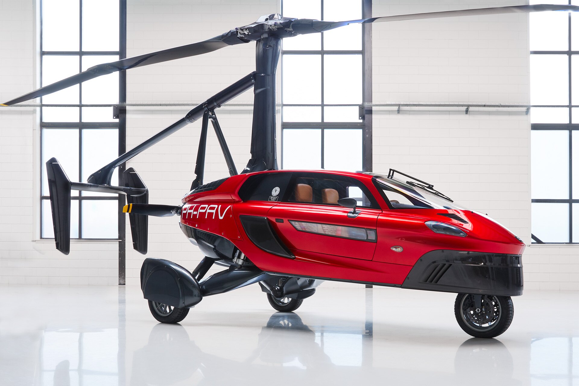 Zdjęcie przedstawia nowoczesny, dwuosobowy pojazd elektryczny w kolorze czerwonym, stojący w sterylnie białej hali. Korpus pojazdu przypomina mały bolid na trzech kołach, jednym przednim i dwóch tylnych. Na stelażu, którego wsporniki są umieszczone na tyle pojazdu i na dachu, widoczne jest śmigło. 