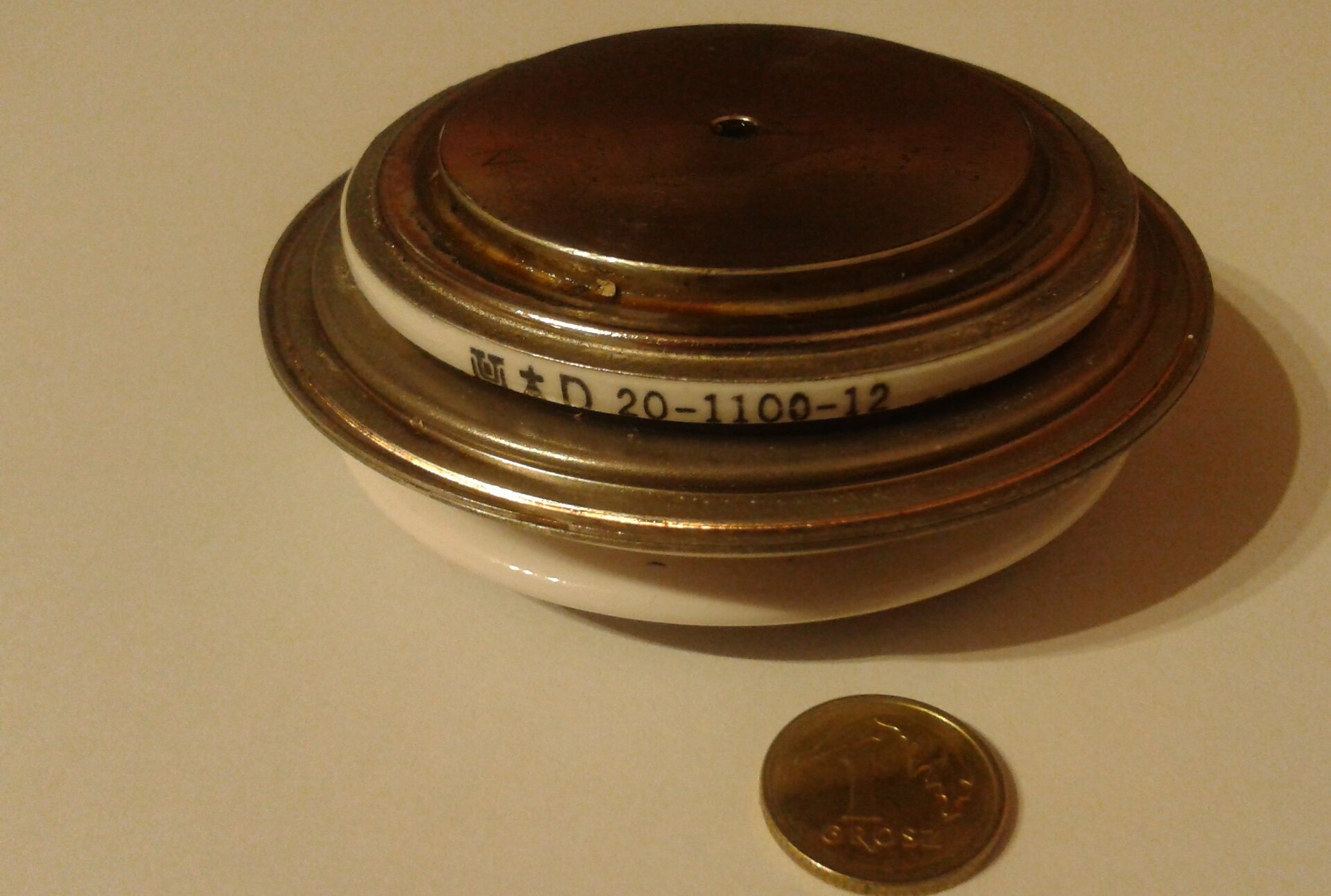 Rys. 2. Zdjęcie przedstawia diodę prostowniczą szybką. Na jasnym tle blatu ułożono krążek w kolorze miedzianym z elementami kremowymi. Krążek składa się z trzech współśrodkowych dysków ułożonych w płaszczyźnie poziomej. Najmniejszą średnicę ma dysk górny, natomiast dysk dolny największą. Obok diody prostowniczej położono monetę jednogroszową. Moneta jest kilkukrotnie mniejsza niż dioda.