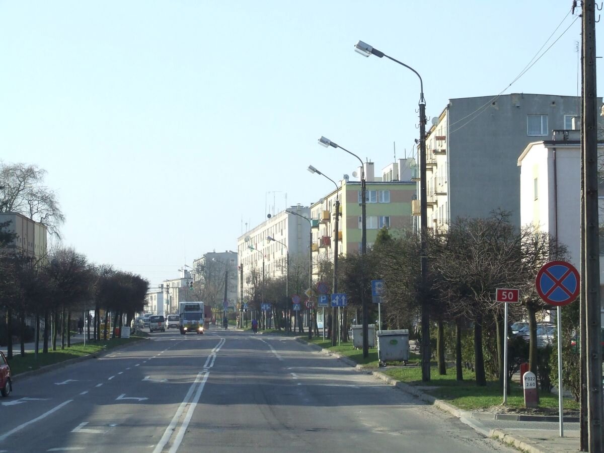 Zdjęcie przedstawia Ciechanów. Na środku znajduje się droga, po której jedzie ciężarówka oraz samochody osobowe. Po obu stronach drogi rosną drzewa i trawa oraz stoją latarnie. Za nimi znajdują się bloki mieszkalne.