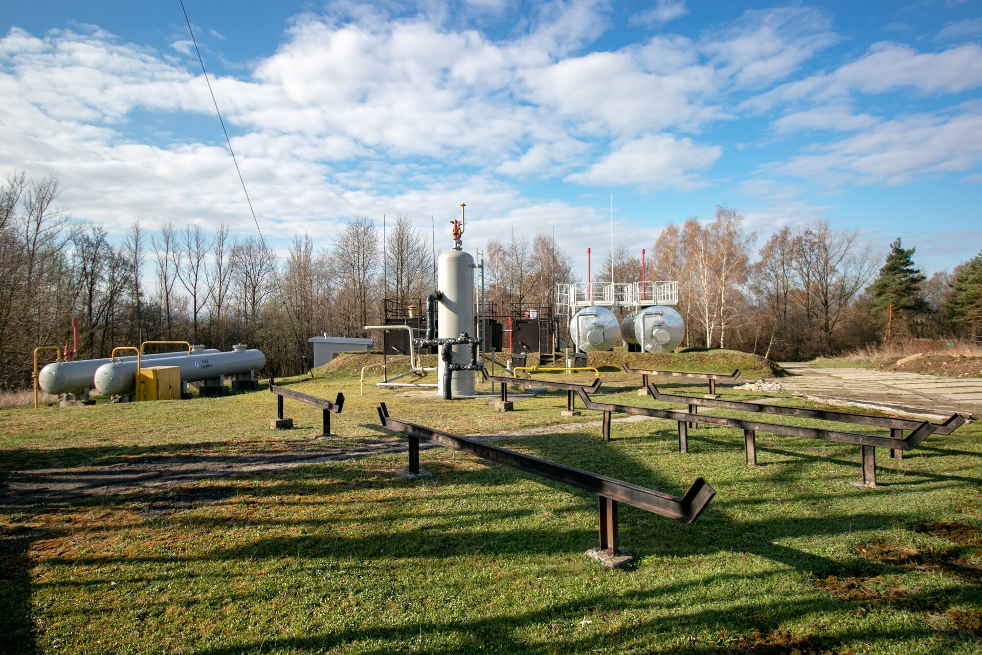 Na zdjęciu widać napowierzchniowe urządzenia do eksploatacji ropy naftowej i gazu ziemnego z widoczną rampą rurową, separatorem dwufaozwym, dwoma zbiornikami cylindrycznymi leżącymi o pojemności sześćdziesięciu metrów sześciennych oraz zbiorniki buforowe gazu ziemnego.W centralnej części zdjęcia znajduje się stalowy separator dwufazowy pionowy typu Rdziesięć łamane na tysiąc, który wykorzystywany jest do rozdziału fazy gazowej od ciekłej. U góry separatora znajduje się czerwony zawór bezpieczeństwa w kształcie odwróconej fajki skierowanej szyjką do góry. Po prawej stronie separatora znajduje się drabina. Za separatorem znajdują się zbiorniki magazynowe ropy naftowej typu cylindrycznego o pojemności V‑sześćdziesiąt metrów sześciennych w obwałowaniu oraz typu skrzyniowego o pojemności V‑pięćdziesiąt metrów sześciennych. Po prawej stronie zdjęcia znajdują się dwa srebrne zbiorniki w kształcie cylindra. Nad zbiornikami znajdują się stalowe pomosty wraz z poręczą zabezpieczającą. Po prawej stronie znajdują się trzy zielone zbiorniki skrzyniowe. Na górze zbiorników znajduje się poręcz zabezpieczająca.. Po lewej stronie zdjęcia znajdują się dwa srebrne, stalowe zbiorniki buforowe w kształcie cygara. Zbiorniki osadzone są na metalowych nóżkach, które znajdują się na podmurówce. Do góry zbiorników przyłączone są żółte rury. Przed pierwszym zbiornikiem znajduje się żółta skrzynia. W tle zdjęcia widać drzewa.