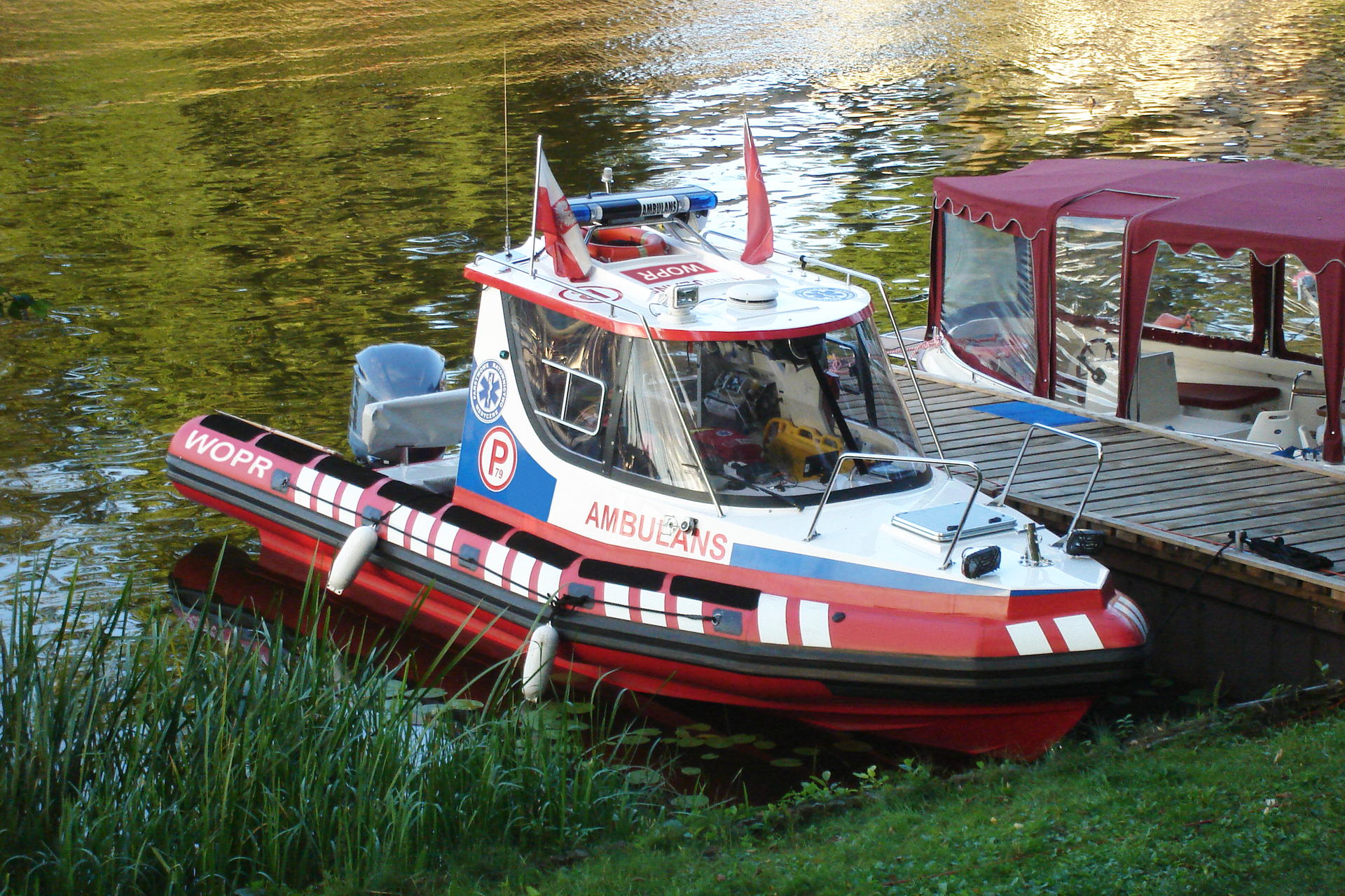 Drugie zdjęcie na prawo to mała łódź ratunkowa WOPR. Łódź przycumowana do brzegu. Kabina kapitana oszklona. Za kabiną dwa miejsca siedzące. Na dachu kabiny dwie biało-czerwone flagi.
