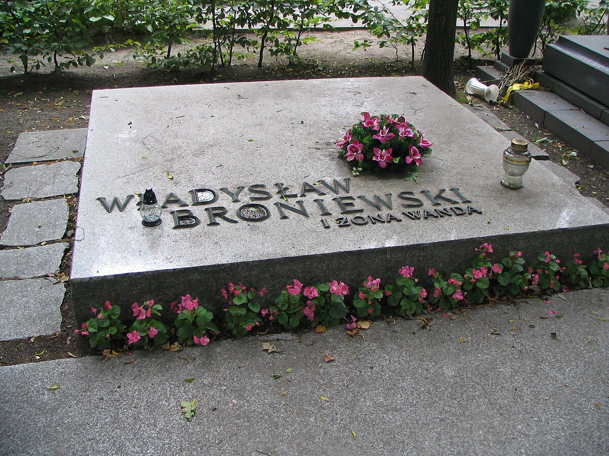 Zdjęcie przedstawia kamienną, kwadratową płytę nagrobną z napisem: Władysław Broniewski i żona Wanda. Na płycie stoją dwa znicze oraz leży mały wieniec z fioletowych kwiatów. Przed płytą rośnie rząd drobnych, różowych kwiatków. Nagrobek otoczony jest chodnikiem ułożonym z kamiennych płyt. W tle widać pień drzewa i krzewy. Po prawej stronie - kolejny nagrobek, a obok niego przewrócony wazon i suche kwiaty.