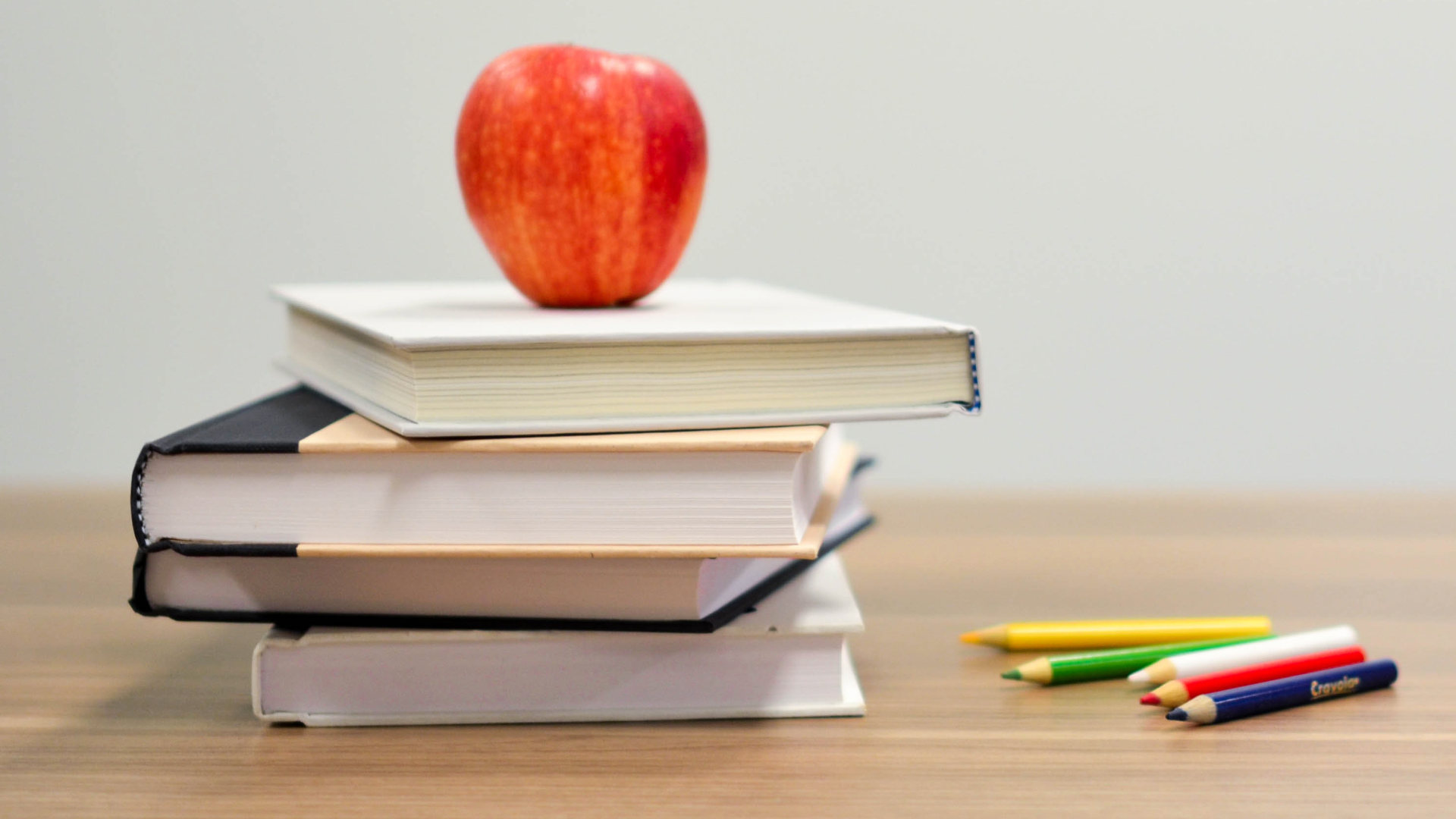Na zdjęciu widocznych jest kilka książek. Książki leżą nierówno, jedna na drugiej. Na książkach znajduje się jabłko. Obok książek, na stole leży kilka kolorowych kredek ołówkowych.