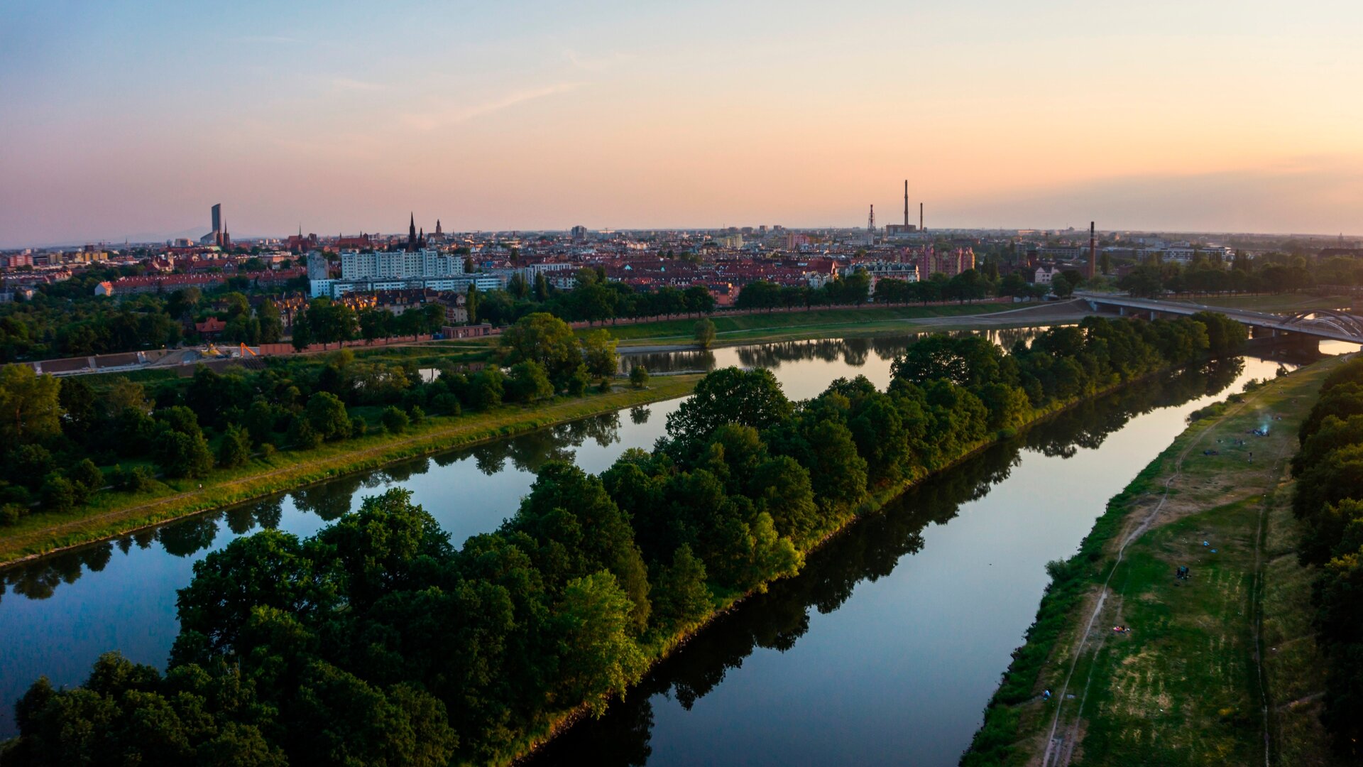 Fotografia przedstawiająca widok kanałów Odry we Wrocławiu. Trzy kanały poprzecinane pasami zieleni zbiegają się z prawej strony zdjęcia. Po tej samej stronie widoczny most. W tle panorama miasta.
