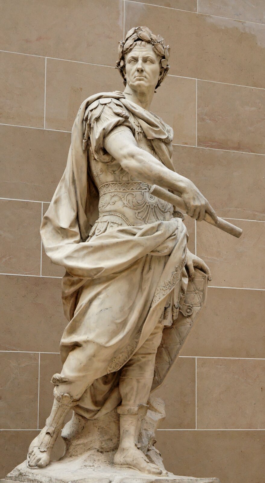Ilustracja przedstawia rzeźbę „Juliusz Cezar” autorstwa Nicolasa Coustou. Rzeźba ukazuje sylwetkę mężczyzny - Juliusza Cezara. Postać mężczyzny stoi odziany w rzymskie szaty, na ramiona zarzuconą ma pelerynę. Postać na głowę założony ma laurowy wianek. W prawej dłoni trzyma papirus, lewą dłoń opiera o tarczę postawioną za nim. 