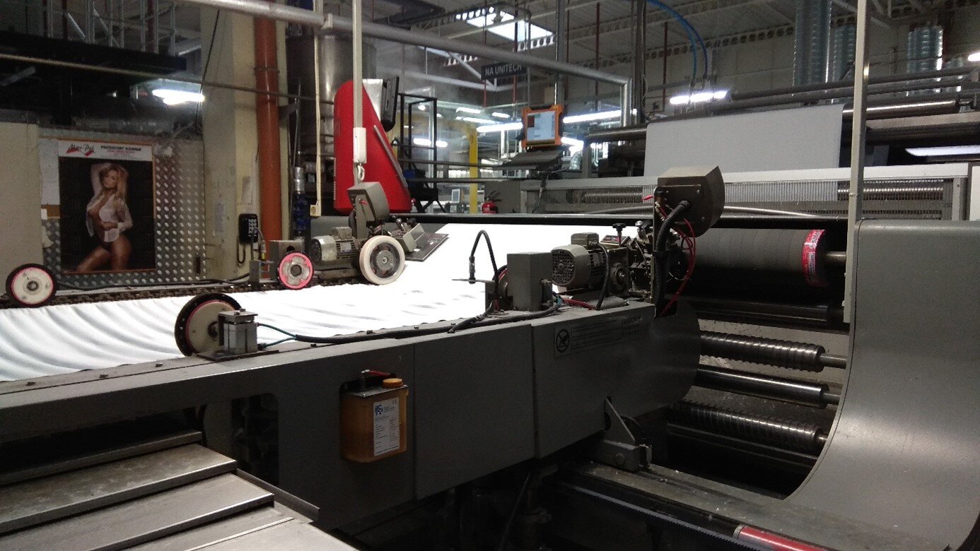 Zdjęcie przedstawia stabilizator stojący w hali produkcyjnej. Urządzenie składa się z taśmy transportującej biały materiał oraz z kilku wałów, przez który przechodzi materiał.