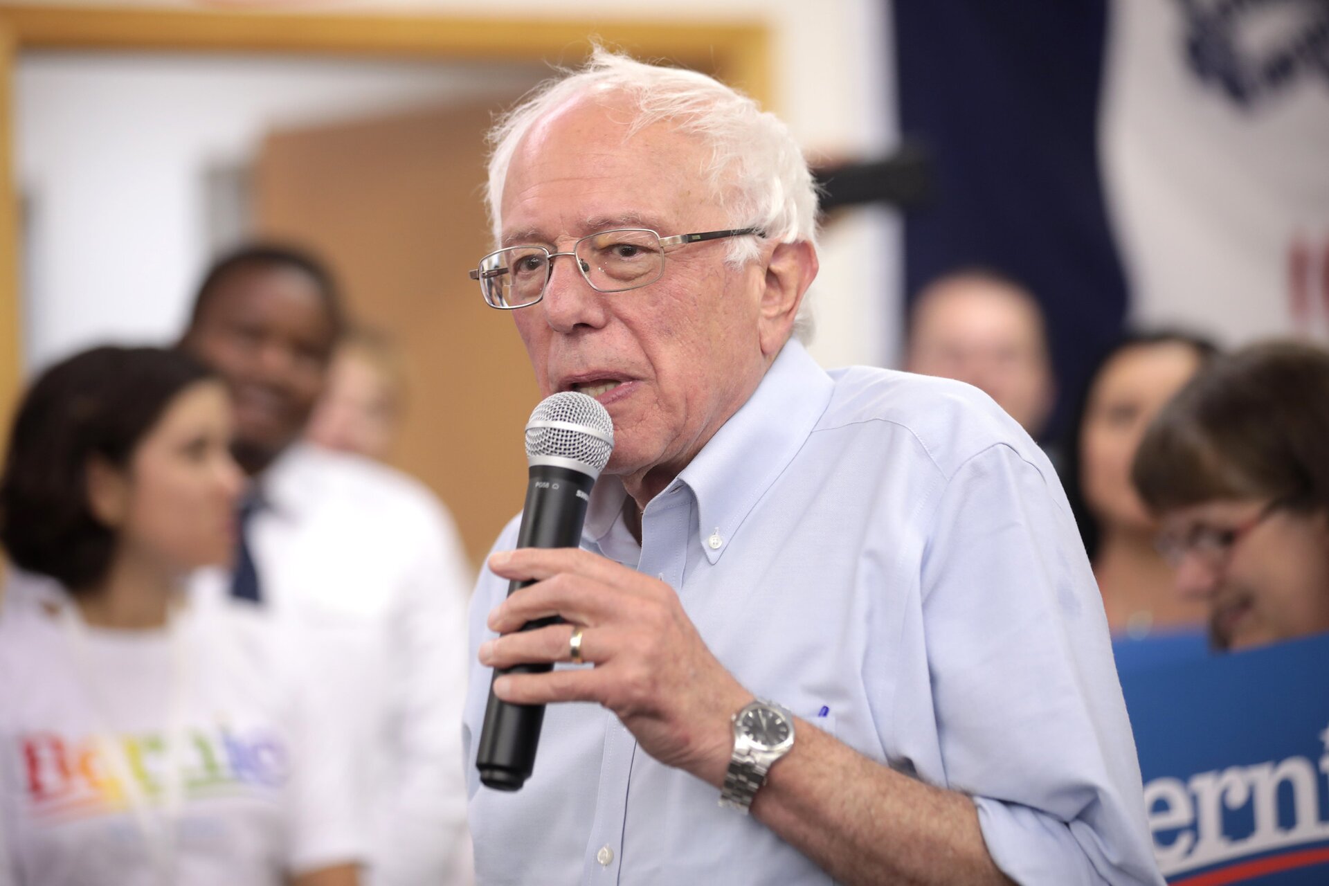 Zdjęcie przedstawia starszego, siwego mężczyznę. Mężczyzna ma pociągła twarz z widocznymi zmarszczkami. Ma założone okulary. Trzyma w ręku mikrofon, przemawia. Za jego plecami, na drugim planie, widać młodych ludzi.