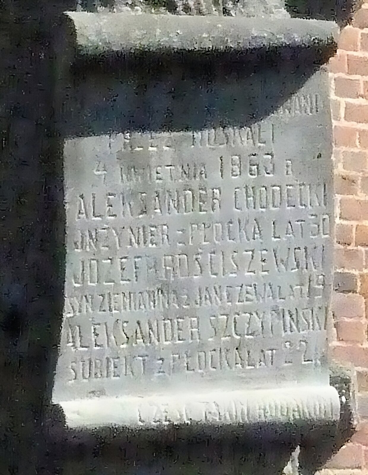 Zdjęcie przedstawia tablicę pamiątkową umieszczoną na ceglanej ścianie. Część napisów nie jest czytelna. Zdjęcie zostało wykonane w świetle dziennym.