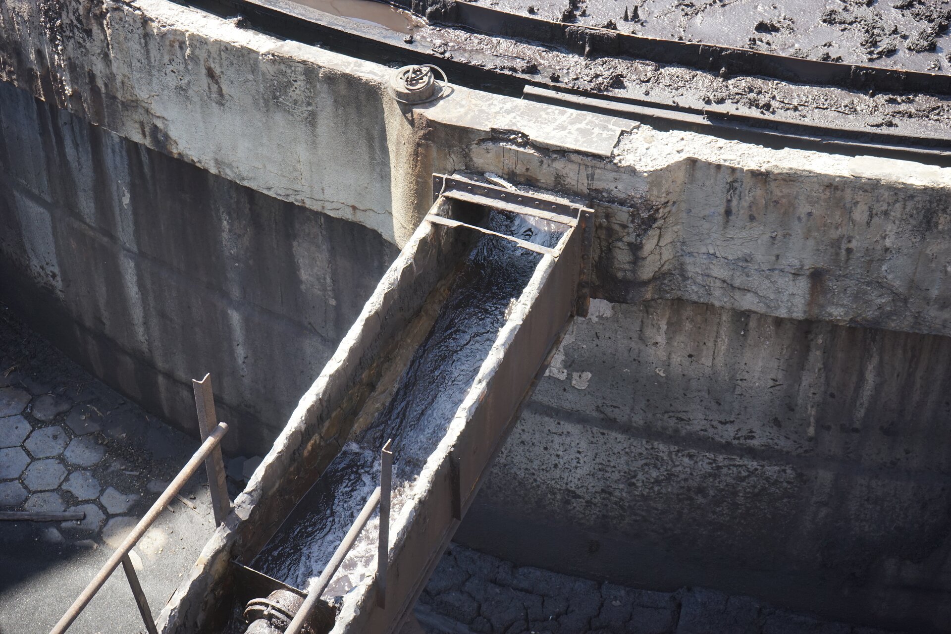 Zdjęcie przedstawia odpływ cieczy klarownej z zagęszczacza promieniowego. Fragment pierścieniowatego przelewu zbiornika głównego zagęszczacza zaopatruję odpływ w ciecz. Rynna przelewowa po jednej stronie zbiornika połączona jest z odpływem cieczy klarownej. Odpływ na zdjęciu poprowadzony jest o łuku rynny w górnej części. ku lewemu dolnemu rogowi fotografii. Poniżej zdjęcia znajduje się przycisk multimedialny z nagraniem audio. Treść nagrania jest zgodna z tytułem zdjęcia.