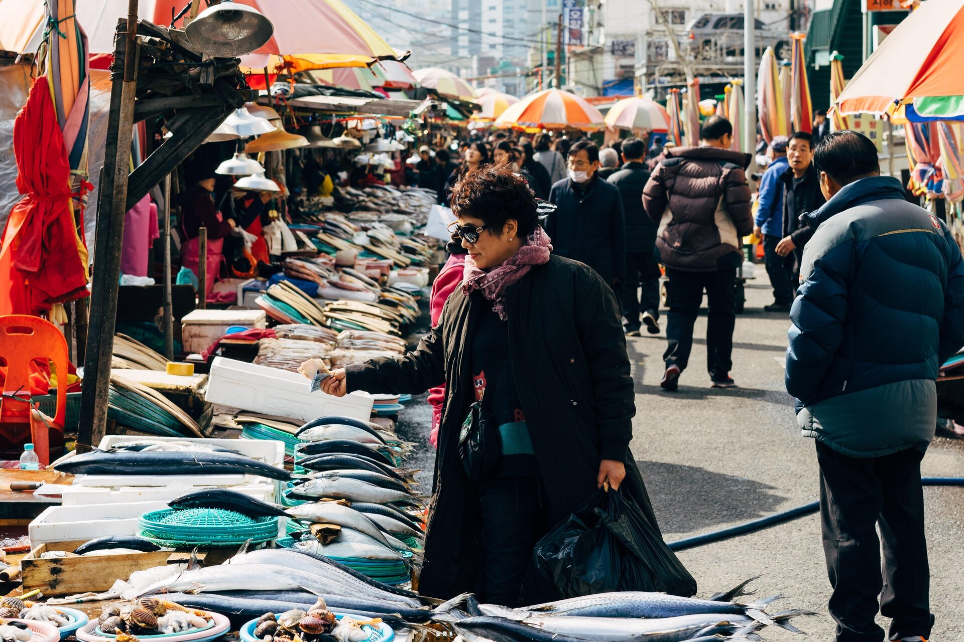 Zdjęcie przedstawia targ rybny na jednej z ulic miasta. Po lewej i prawej stronie uliczki widoczne są stoiska z różnymi produktami, nad którymi rozłożone są kolorowe parasole. Na stoiskach znajdują się ryby, owoce morza, zamknięte i otwarte pojemniki styropianowe oraz kolorowe kosze. Po uliczce w różnych kierunkach przemieszczają się ludzie, z których część robi zakupy. Na pierwszym planie, po lewej stronie, widoczna jest kobieta zwrócona w lewo, która w prawej, wyprostowanej ręce trzyma plik banknotów.