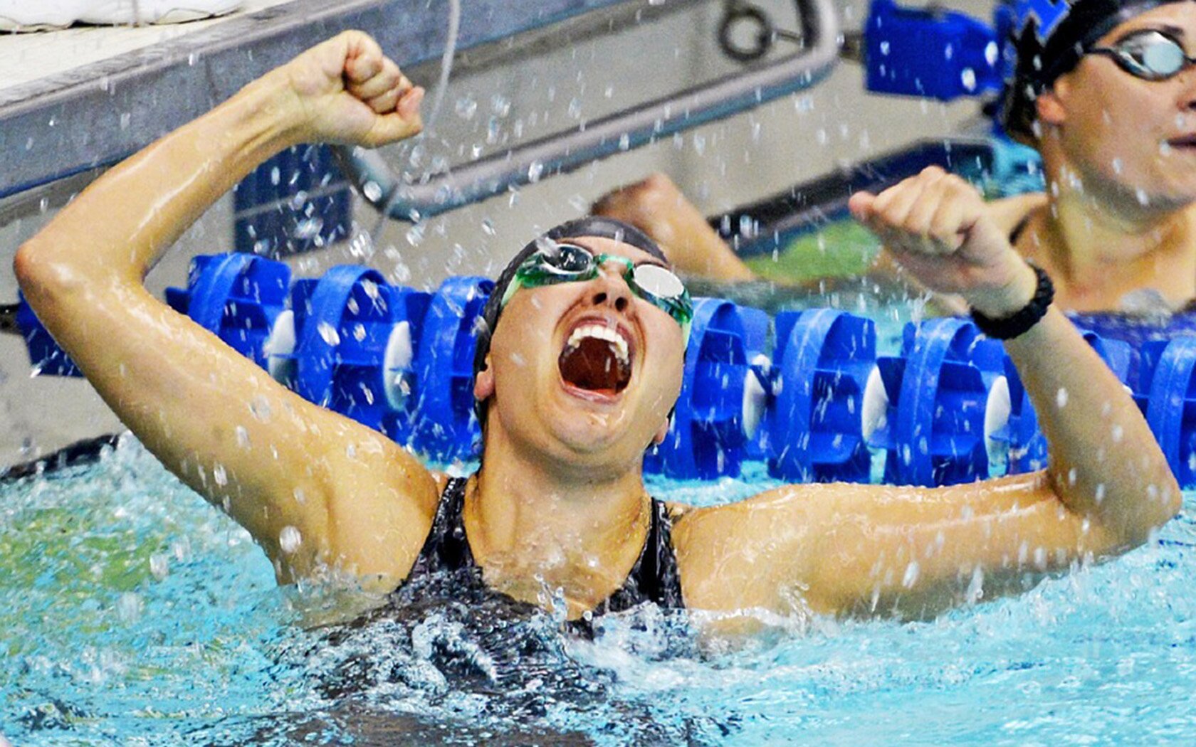 Zdjęcie przedstawia kobietę w stroju pływacki, w basenie. Kobieta zaciskając pięści unosi obie ręce ku górze, a jej buzia jest szeroko otwarta.