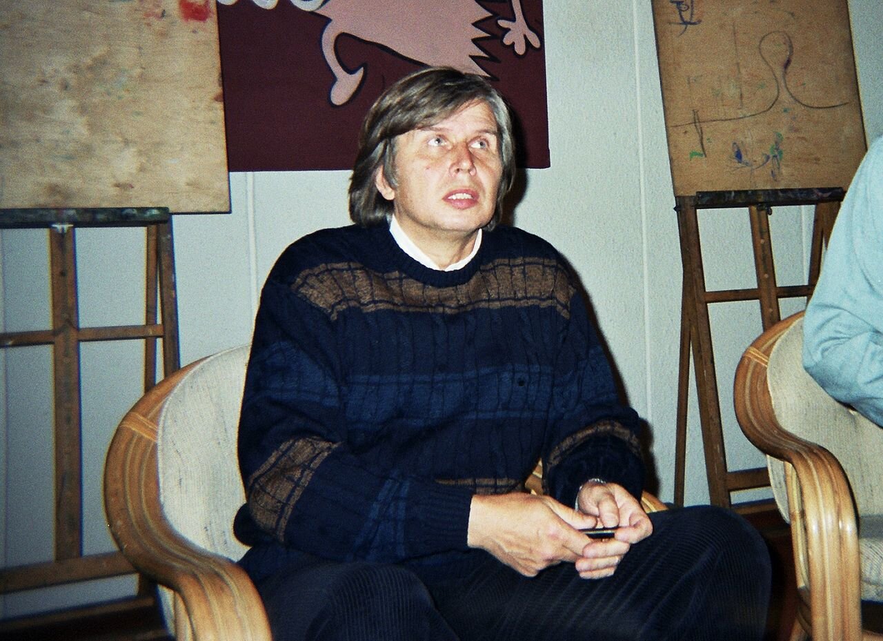 Kolorowe zdjęcie przedstawia mężczyznę w średnim wieku. Mężczyzna ma dłuższe włosy z grzywką i baki. Ubrany jest w granatowy sweter w pasy, spod którego wystaje kołnierzyk białej koszuli. Siedzi w fotelu. Za nim znajdują się sztalugi. Na ścianie wisi grafika.