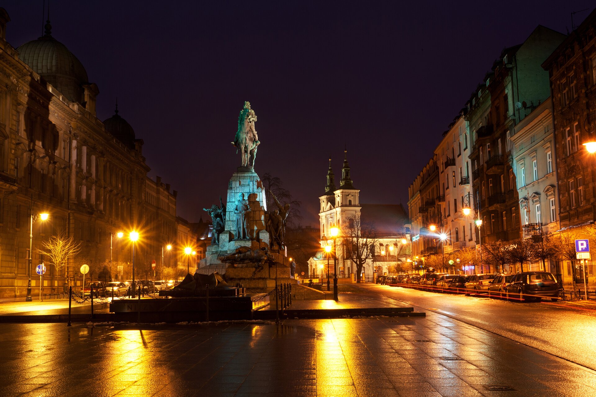 Ilustracja przedstawia pomnik Grunwaldzki w Krakowie. Zdjęcie ukazuje monument w nocnej, miejskiej scenerii. Pomnik oświetlony jest błękitnym światłem. Na pomniku znajduje się postać króla -Władysława Jagiełły na koniu, poniżej postać księcia Witolda.