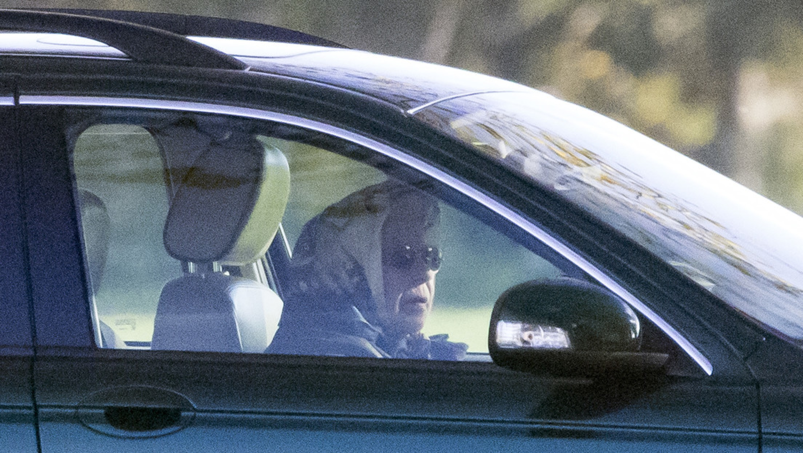 Zdjęcie przedstawia królową Elżbietę II za kierownicą samochodu. Widoczna jest starsza kobieta, która ma zawiązaną na głowie chustkę i założone okulary przeciwsłoneczne. Siedzi za kierownicą ciemnego samochodu.