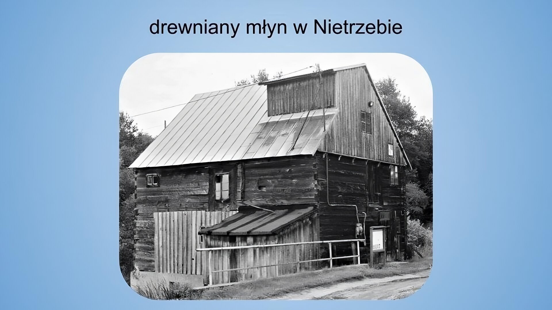 Jasnoniebieskie tło. Na górze napis: drewniany młyn w Nietrzebie. Poniżej czarno‑biała fotografia przedstawiająca drewniany budynek w kształcie sześcianu z niewielką przybudówką i spadzustym dachem. W gónej oraz środkowej części budynku widać trzy niewielkie okna. Przez frontową część elewacji w poziomie i w pionie rozciąga się instalacja elektryczna prowadząca do kabla, który jest rozciągnięty wzdłuż budynku i znika z lewej strony kadru. Na pierwszym planie widoczny jest fragment drogi, a obok niego krótki i niski płot oraz niewielka tablica na dwóch nogach. W tle widoczne są drzewa i jasne niebo.
