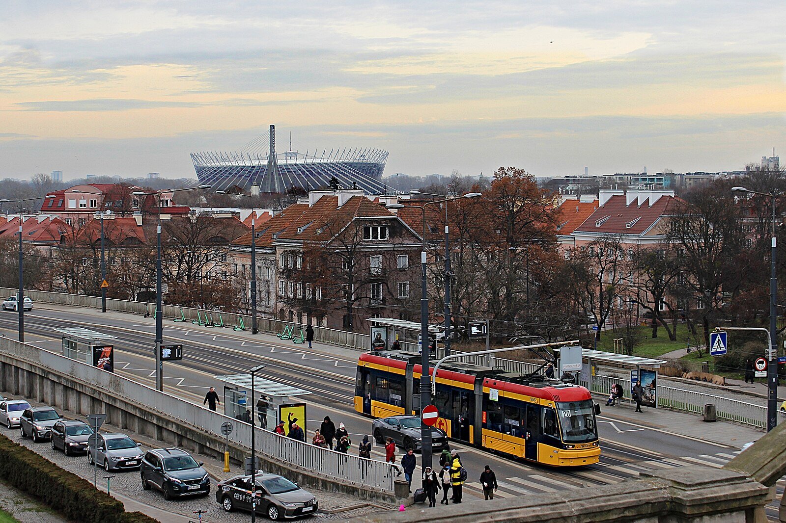 Zdjęcie przedstawia zabytkową zabudowę Warszawy na tle nowoczesnego stadionu. Na pierwszym planie szeroka ulica i tory tramwajowe.
