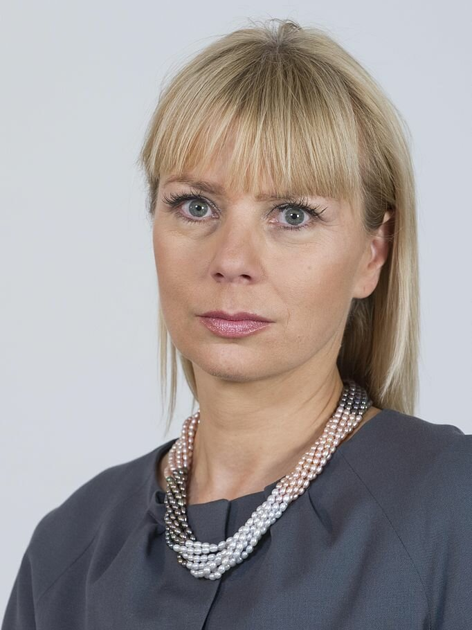 Fotografia przedstawiająca kobietę w sile wieku. Postać siedzi, ukazana jest od piersi w górę, na wprost nas. Kobieta ma owalną twarz, długie włosy z grzywką sięgającą wyrazistych brwi. Ma duże oczy z rzęsami podkreślonymi tuszem i niewielkie zmarszczki wokół oczu. Nos jest prosty, usta uszminkowane. Kobieta jest ubrana w żakiet. Na szyi ma korale ze złączonych sznurów pereł.