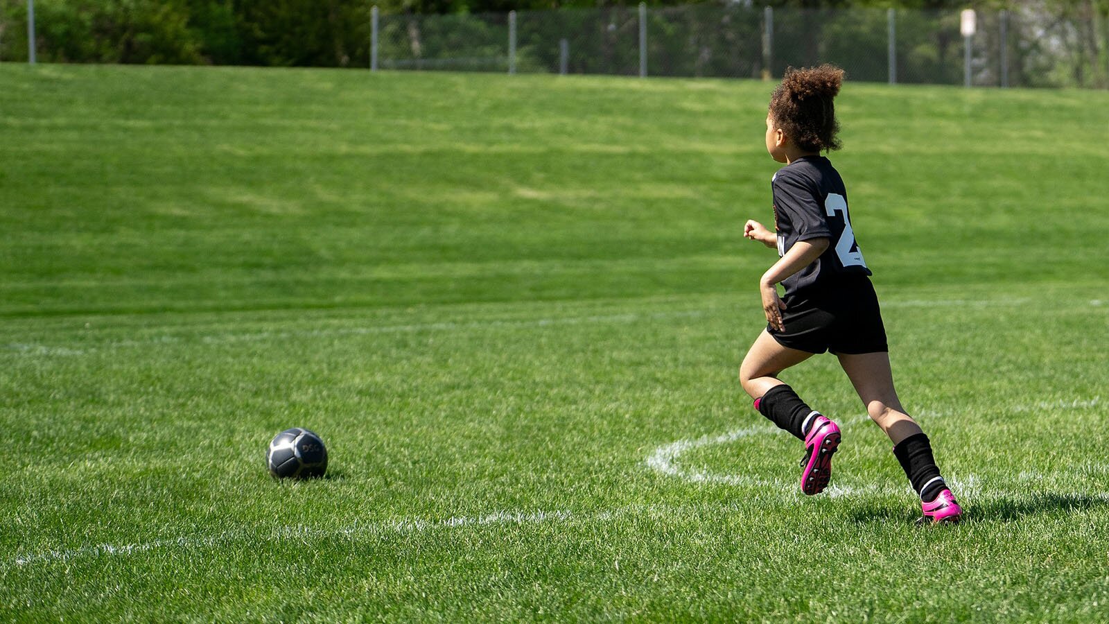 Zdjęcie przedstawia dziewczynkę biegnącą w kierunku piłki leżącej na murawie piłkarskiej. Dziewczyna jest ubrana w strój piłkarski – czarną koszulkę i czarne spodnie i ma lewe kolano podniesione do góry, a prawą nogę wyprostowaną w tyle. 