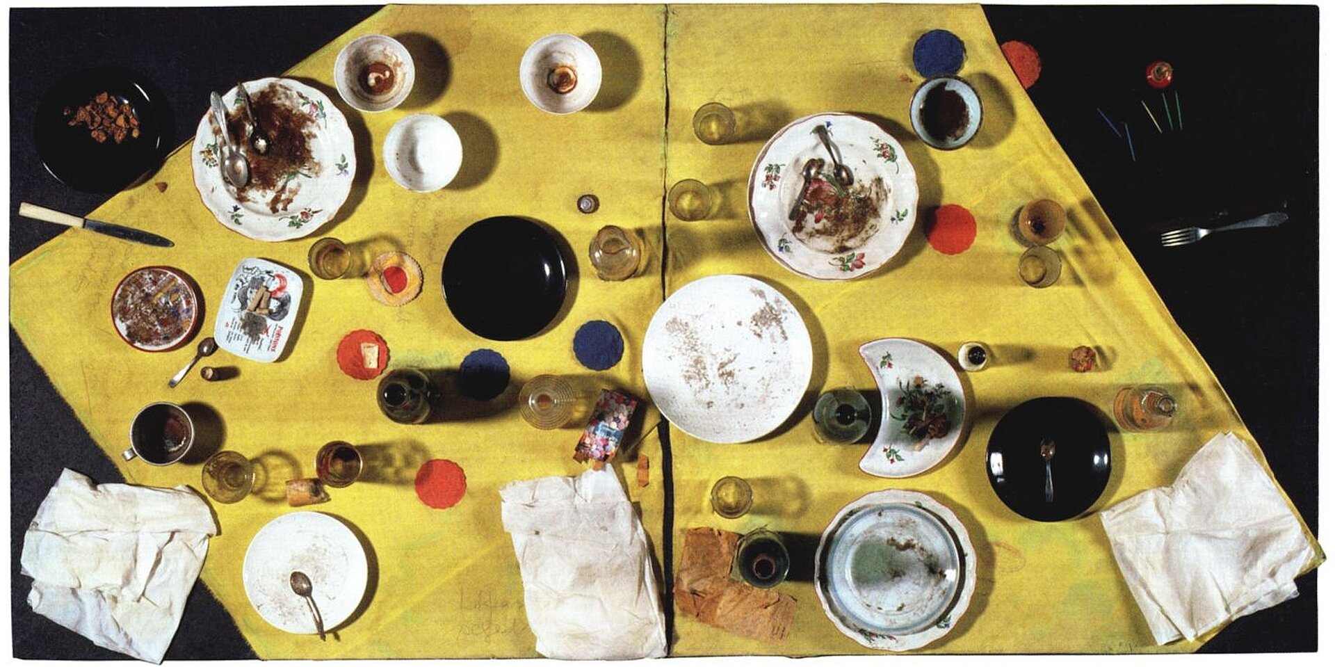 Ilustracja o kształcie poziomego prostokąta pracę  Daniela Spoerriego „Węgierski posiłek”. Ukazuje fotografię wykonaną z góry, przedstawiającą stół nakryty czarnym  obrusem i żółtą serwetą, na której ustawione są brudne naczynia. W szklanych kubkach, butelkach, szklankach i kieliszkach znajdują się resztki płynów, a na talerzykach resztki jedzenia. Widoczne są również zużyte, brudne serwetki. Wszystkie przedmioty rozstawione zostały niedbale. 